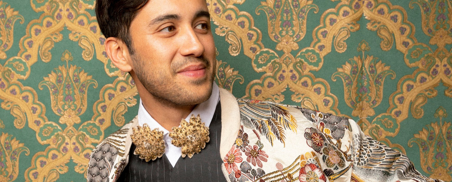 man wearing 2 brooches against a patterned wall drinking an espresso