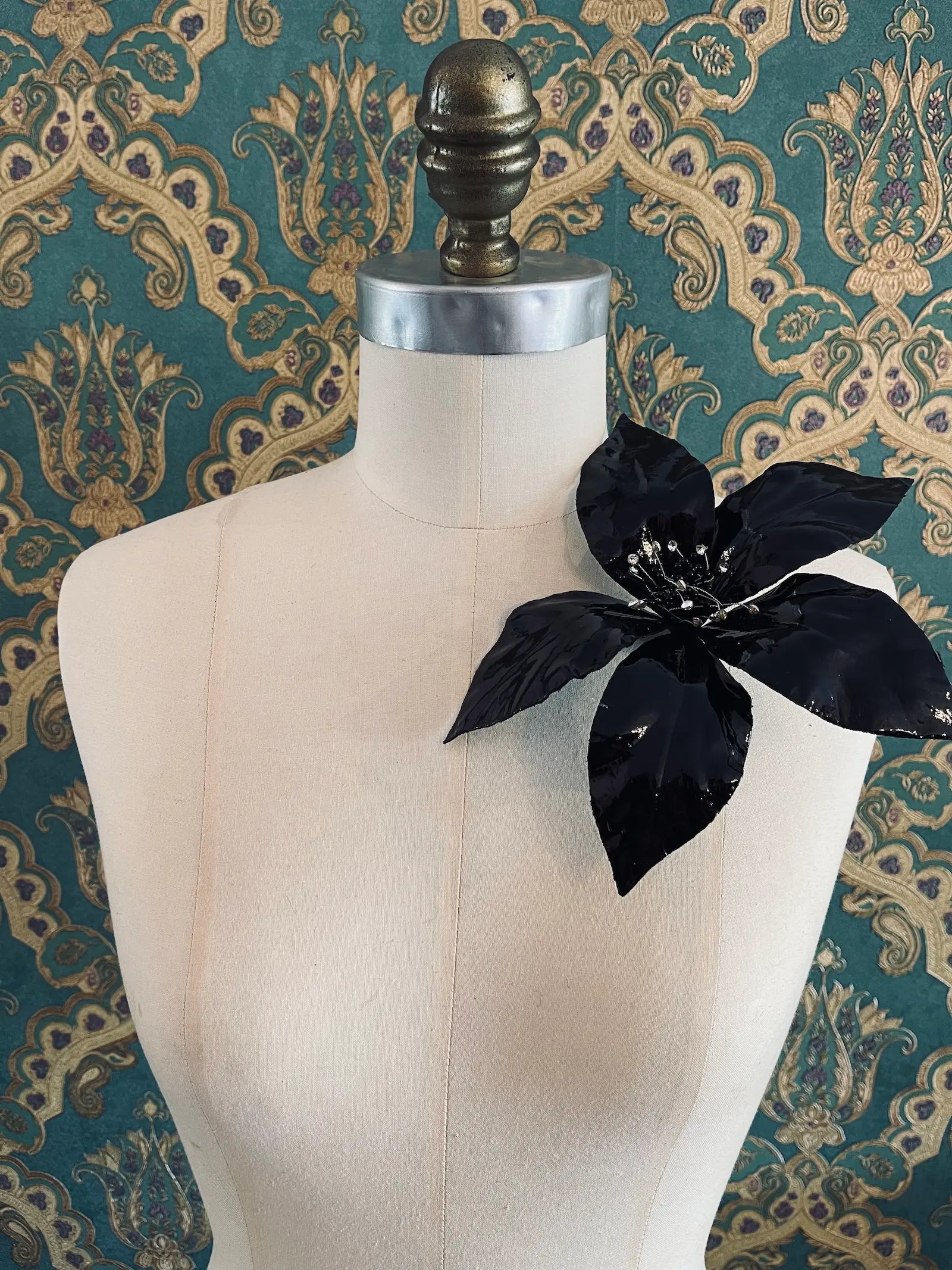 Broochella giglio handmade large patent leather flower in black. Styled on a mannequin. The background is green and gold damask wallpaper