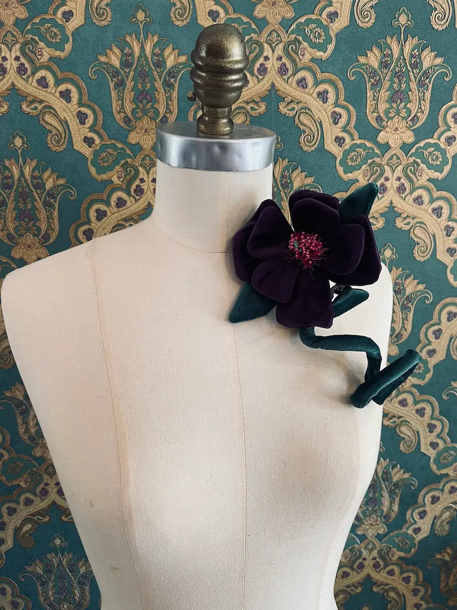 Broochella Velluto Purple and  green handmade velvet flower designer brooch with stem and leaves. Styled on a mannequin. The background is green and gold damask wallpaper