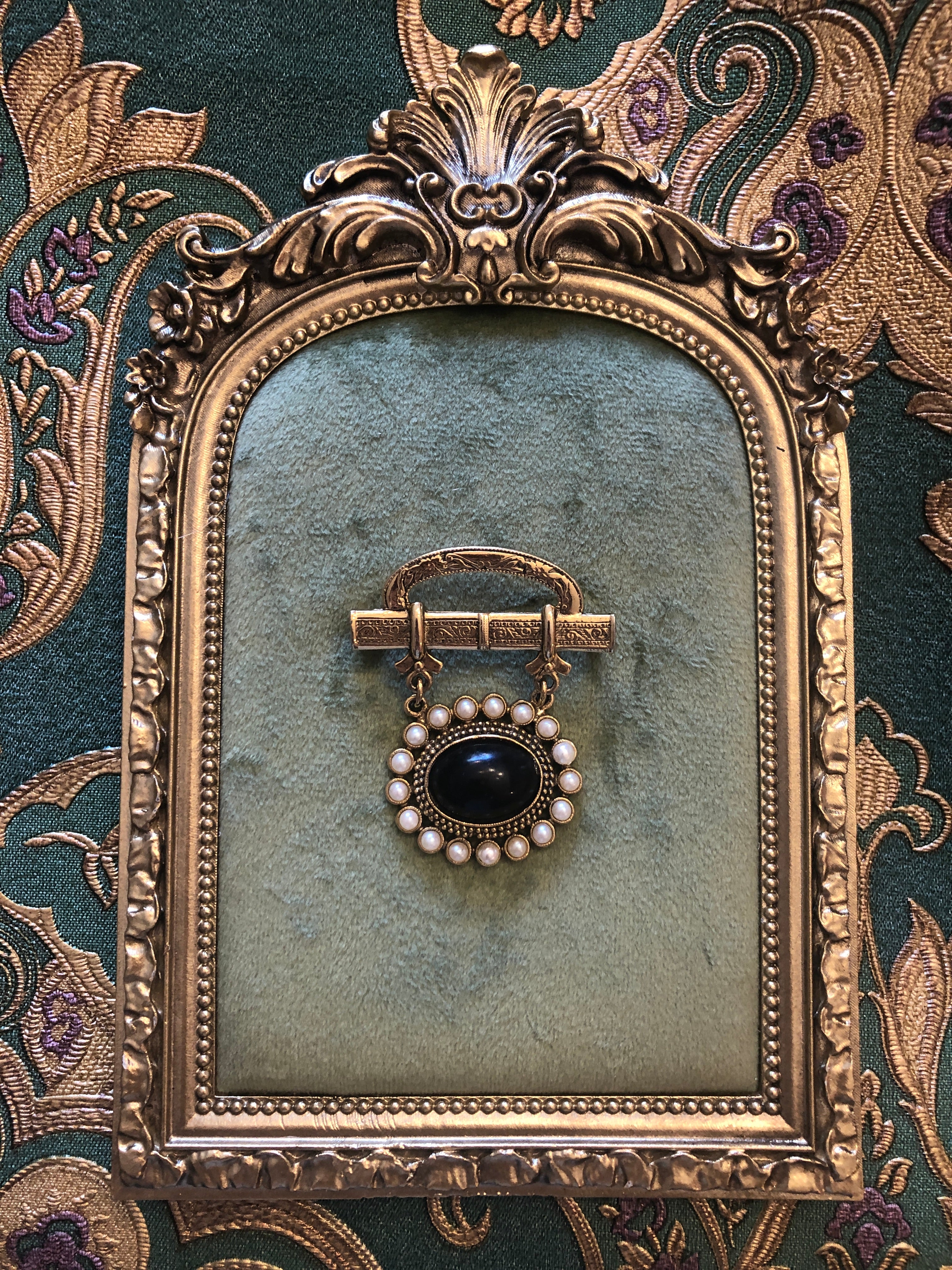 A 5cm x 4cm vintage gold-tone Victorian mourning style brooch featuring an engraved scrollwork bar top with domed oval plaque, suspending via fine chains a circular drop pendant set with a deep black onyx oval cabochon surrounded by a full halo of creamy seed pearls, photographed on sage green velvet.