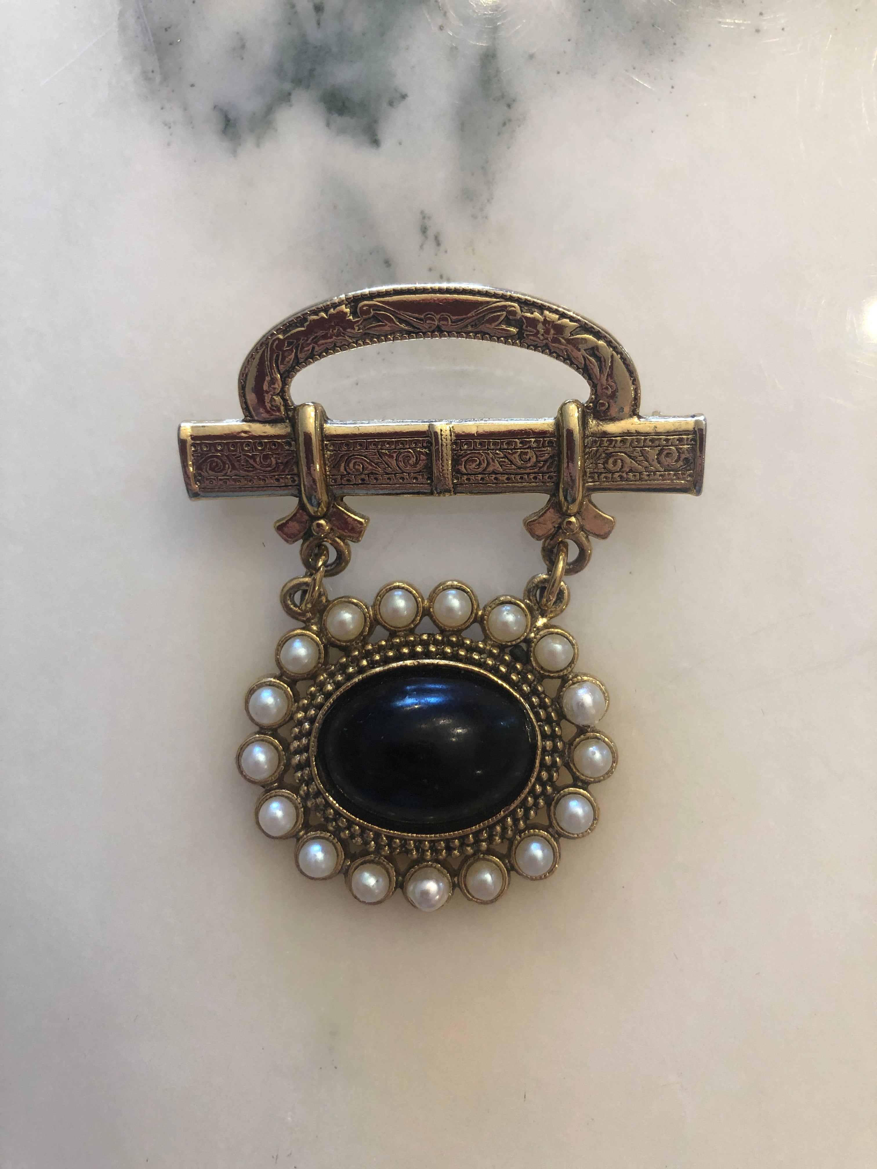 A 5cm x 4cm vintage gold-tone Victorian mourning style brooch featuring an engraved scrollwork bar top with domed oval plaque, suspending via fine chains a circular drop pendant set with a deep black onyx oval cabochon surrounded by a full halo of creamy seed pearls, photographed on marble table