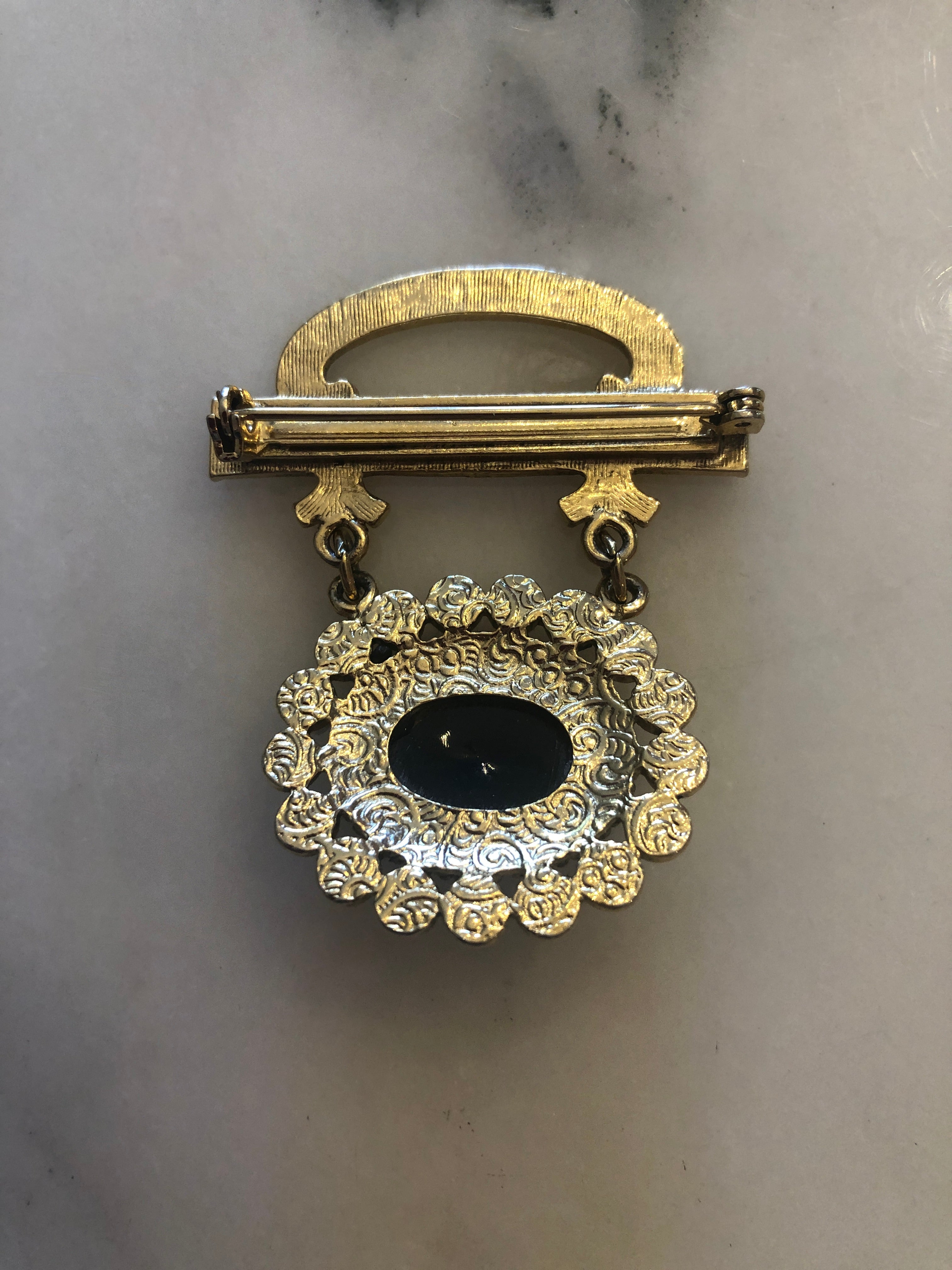 A 5cm x 4cm vintage gold-tone Victorian mourning style brooch featuring an engraved scrollwork bar top with domed oval plaque, suspending via fine chains a circular drop pendant set with a deep black onyx oval cabochon surrounded by a full halo of creamy seed pearls, photographed on marble table