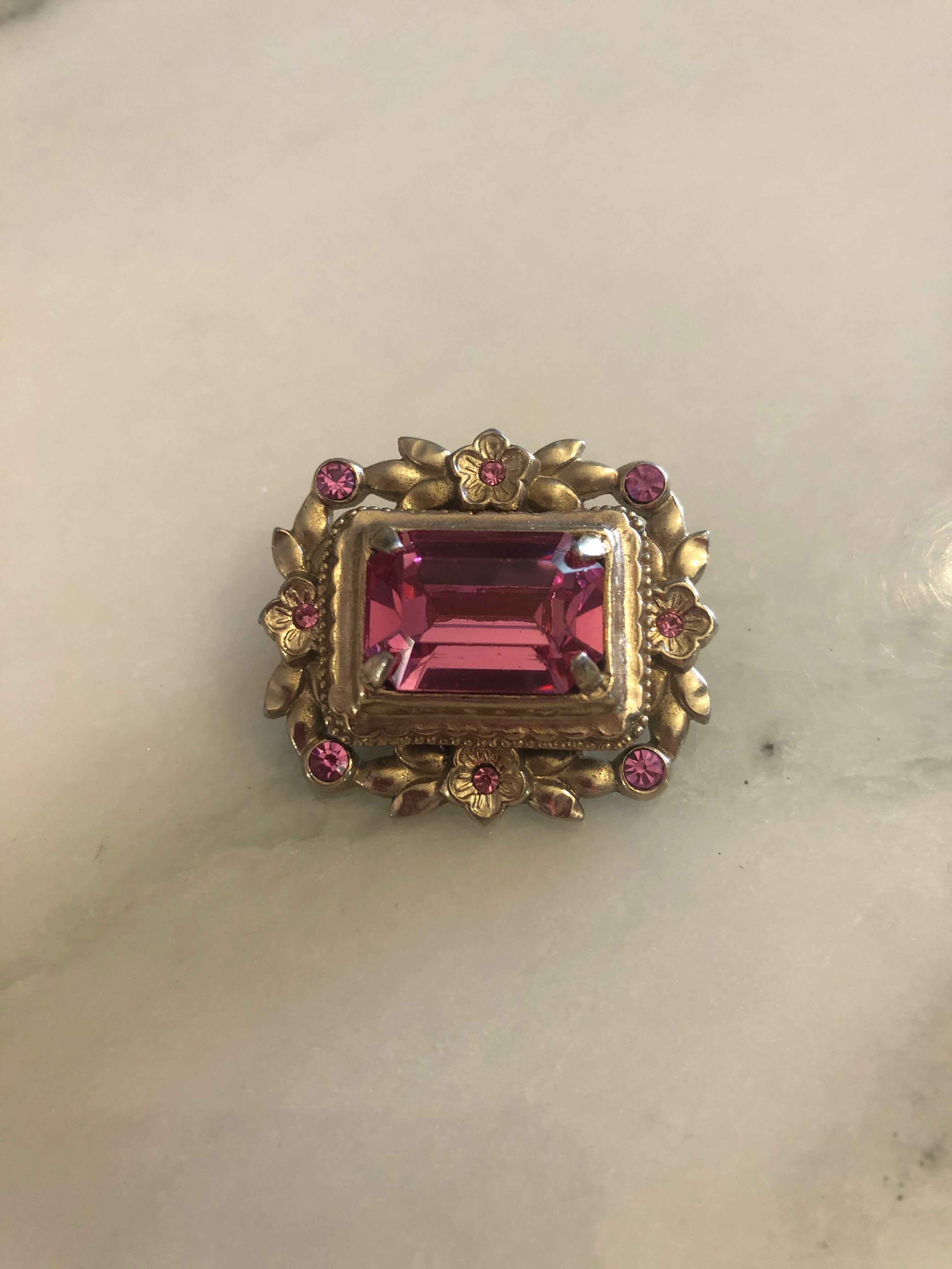 A small vintage gold-tone rectangular brooch featuring a large faceted emerald-cut pink crystal centre stone set within a beaded frame, surrounded by a floral wreath of matte gold-tone flower and leaf motifs with six pink rhinestone accents, photographed on a white marble surface.