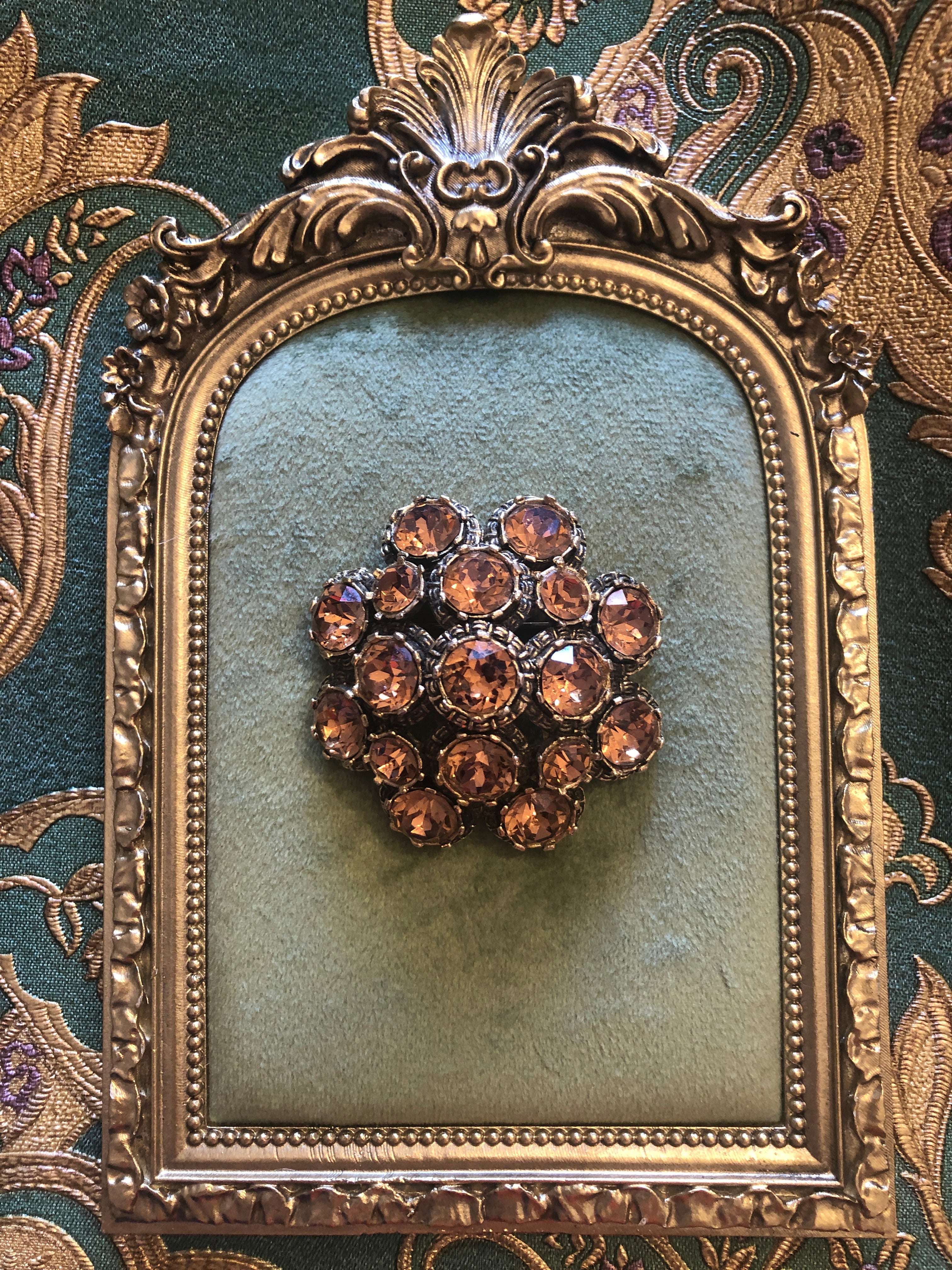 A vintage signed Elizabeth Reimer Éclat d'Ambre Paris silver-tone cluster brooch featuring graduating layers of faceted round champagne and amber crystal rhinestones in ornate individual settings, forming a domed floral cluster arrangement, photographed on a green velvet tray with a gold frame. The background is green and gold damask wallpaper