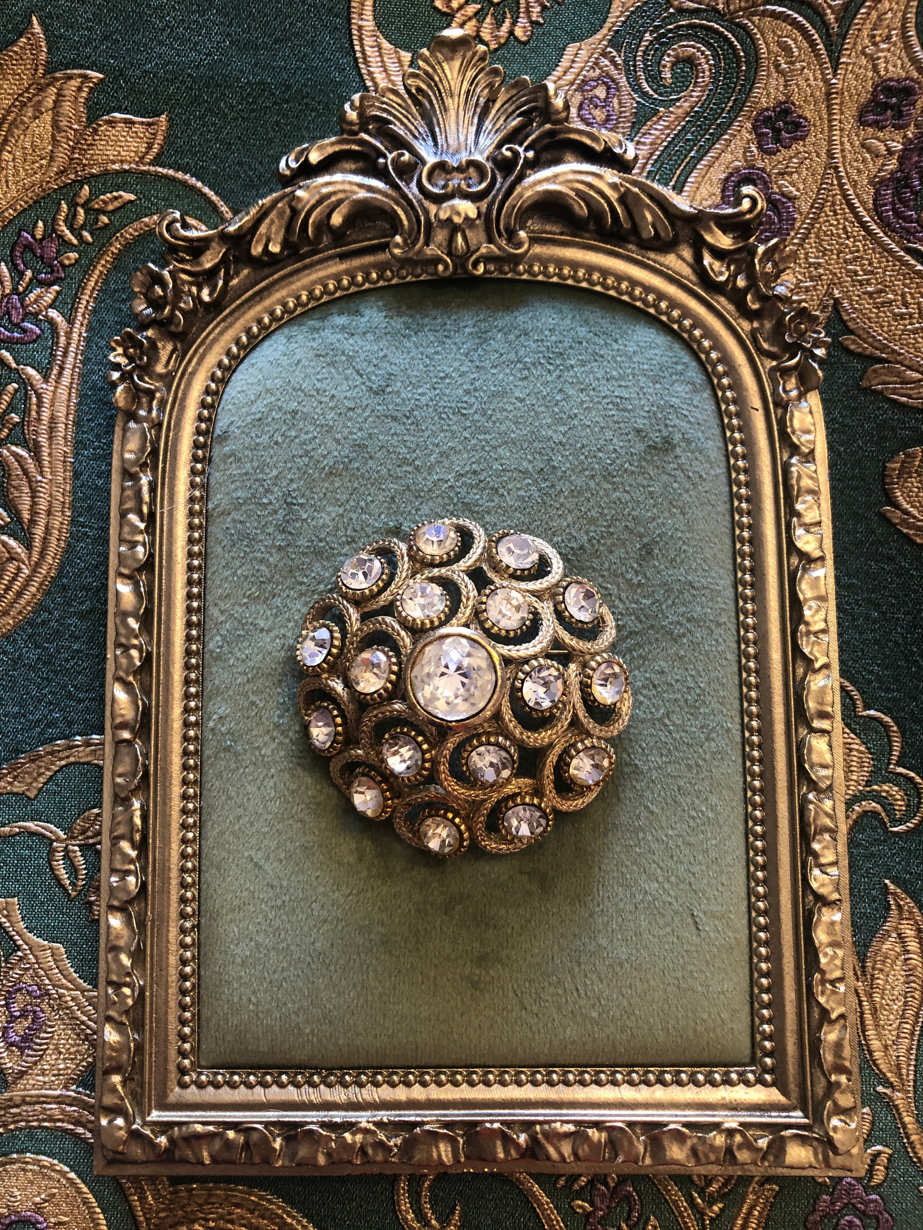 A vintage circular gold-tone brooch featuring a large faceted clear crystal centre stone surrounded by an intricate openwork rope twist scrollwork frame set with fourteen aurora borealis crystal rhinestones in individual beaded settings, photographed on a white marble surface.
