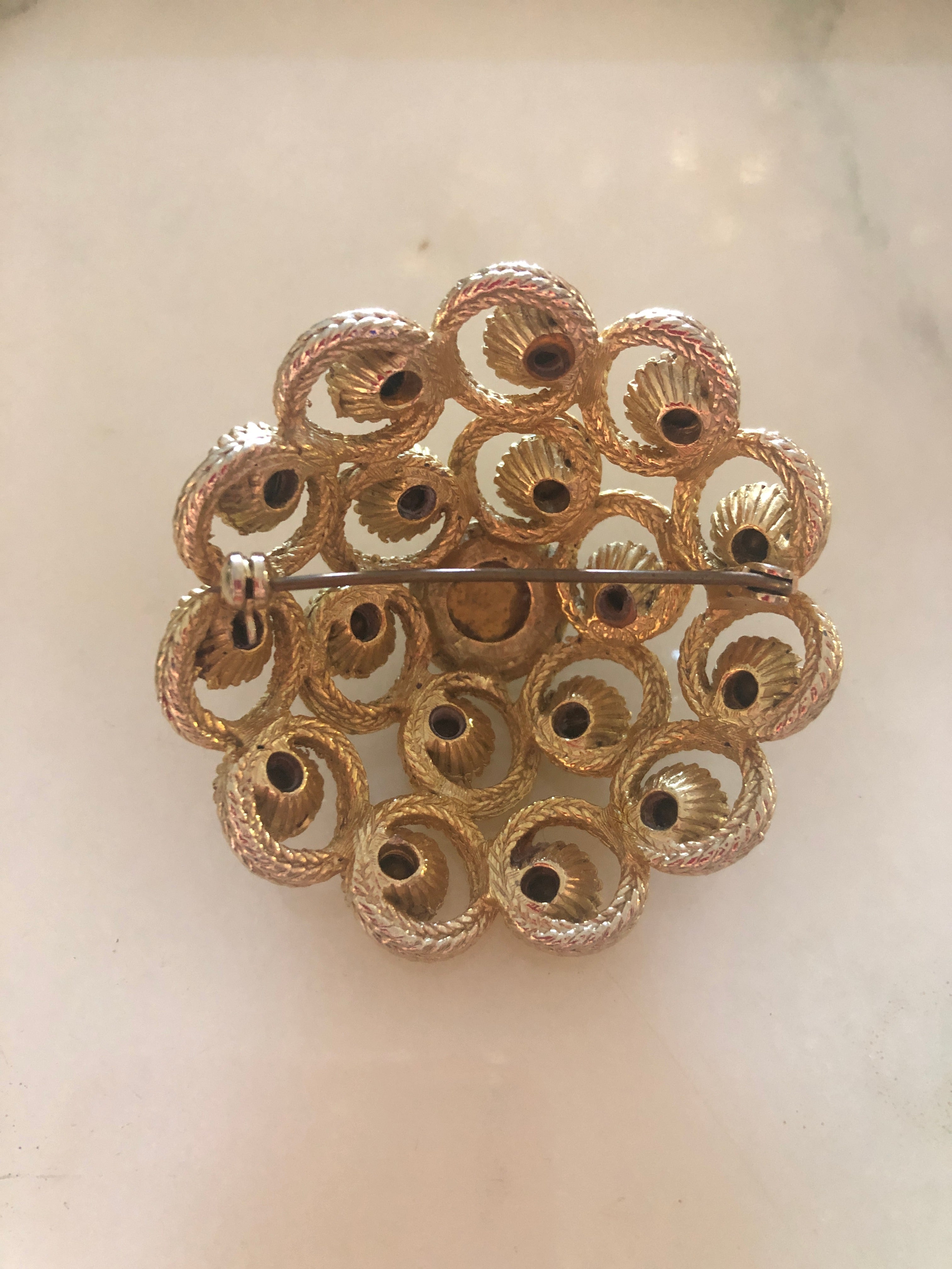 A vintage circular gold-tone brooch featuring a large faceted clear crystal centre stone surrounded by an intricate openwork rope twist scrollwork frame set with fourteen aurora borealis crystal rhinestones in individual beaded settings, photographed on a white marble surface.
