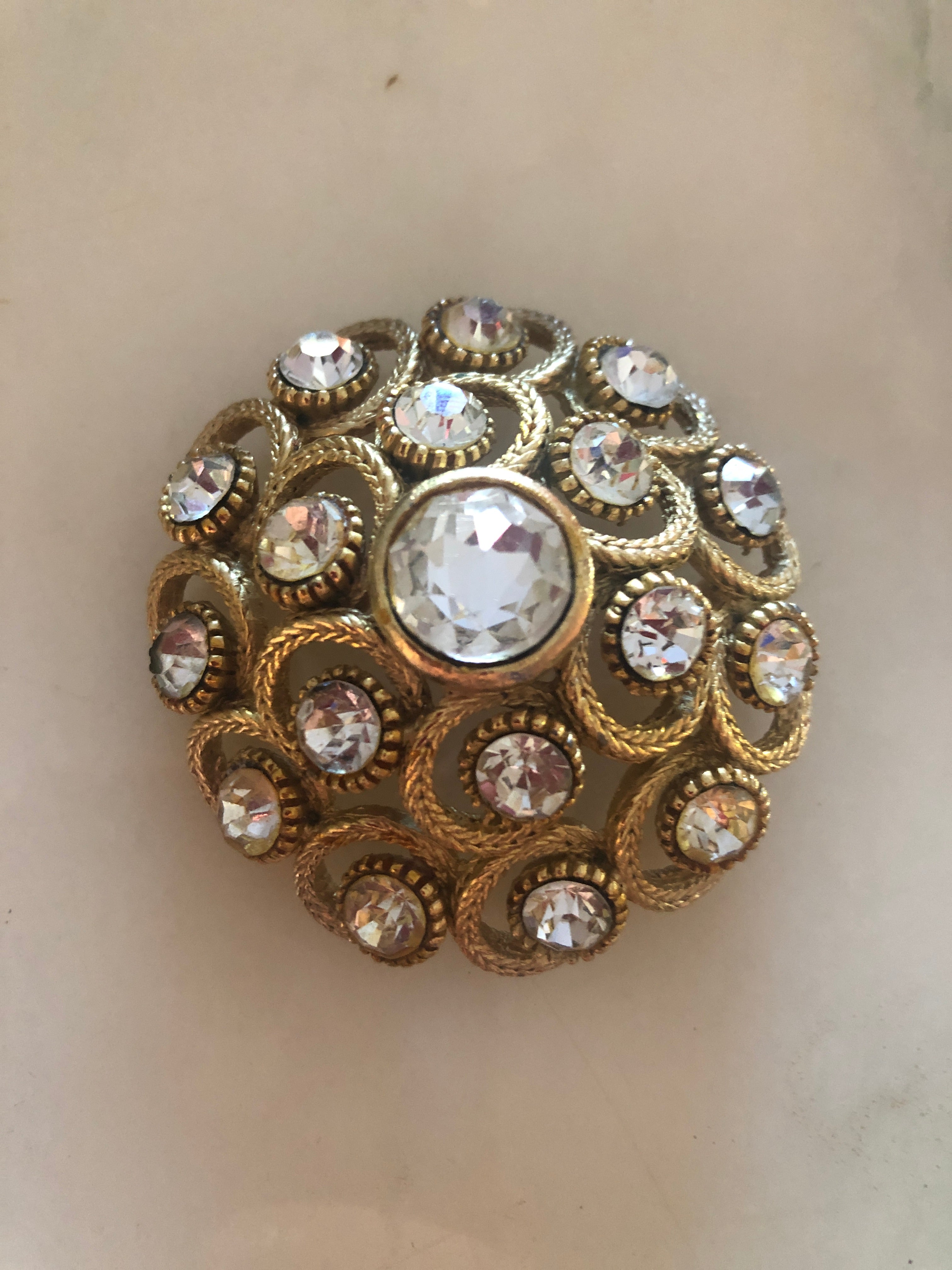 A vintage circular gold-tone brooch featuring a large faceted clear crystal centre stone surrounded by an intricate openwork rope twist scrollwork frame set with fourteen aurora borealis crystal rhinestones in individual beaded settings, photographed on a white marble surface.