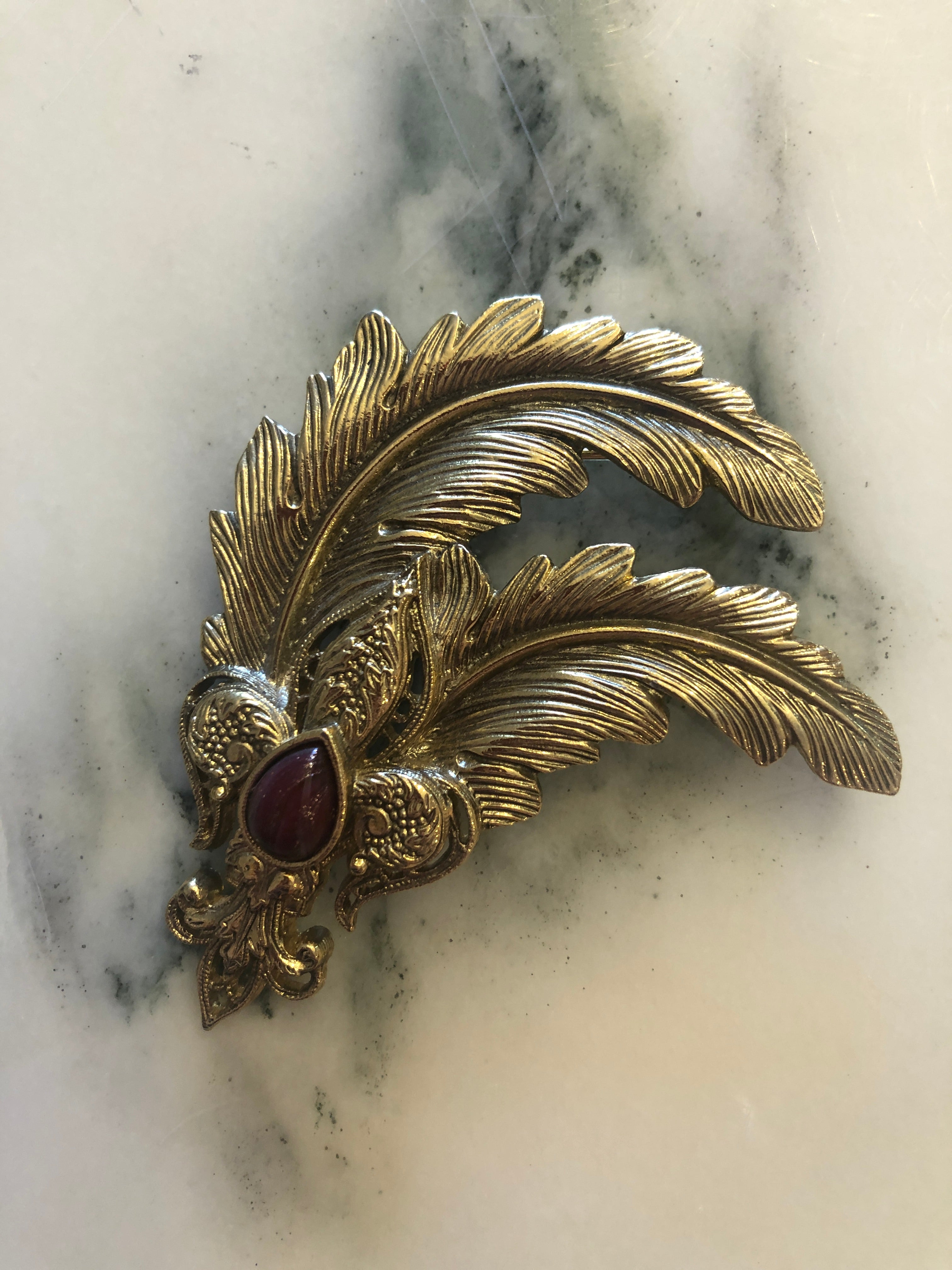 A vintage gold-tone Plume Impériale brooch featuring three sweeping sculpted feather plumes with finely detailed barb texturing, anchored by an ornate repousse scrollwork base set with a deep ruby red pear-cut cabochon stone accent, photographed on a white marble surface.
