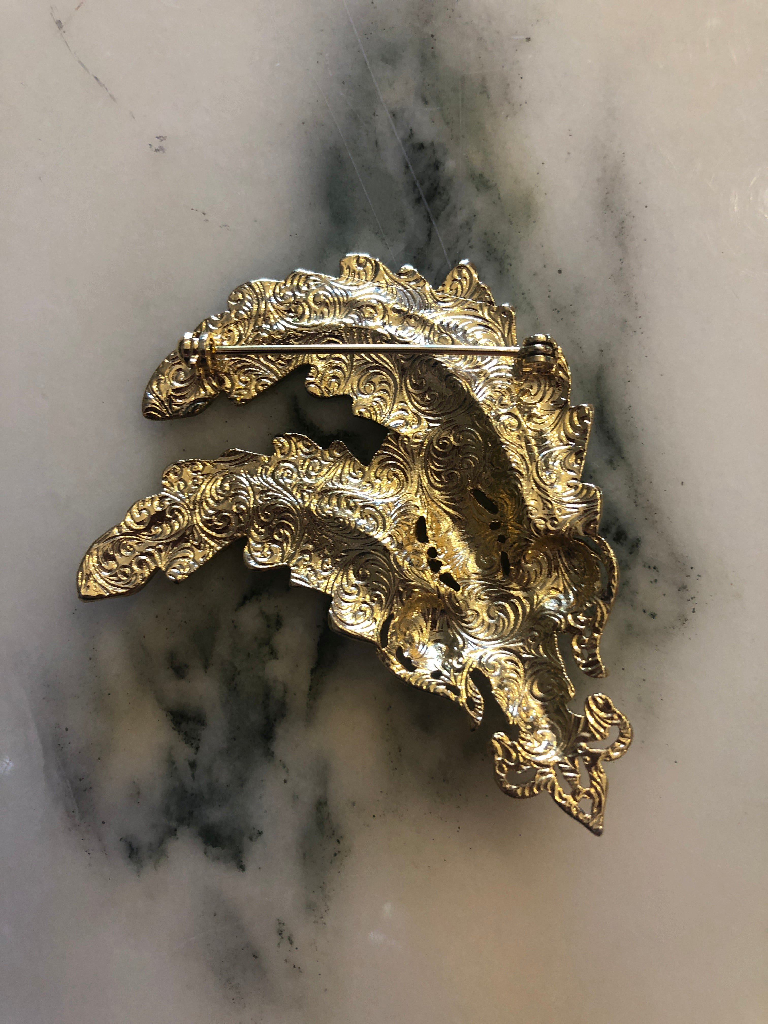 A vintage gold-tone Plume Impériale brooch featuring three sweeping sculpted feather plumes with finely detailed barb texturing, anchored by an ornate repousse scrollwork base set with a deep ruby red pear-cut cabochon stone accent, photographed on a white marble surface.