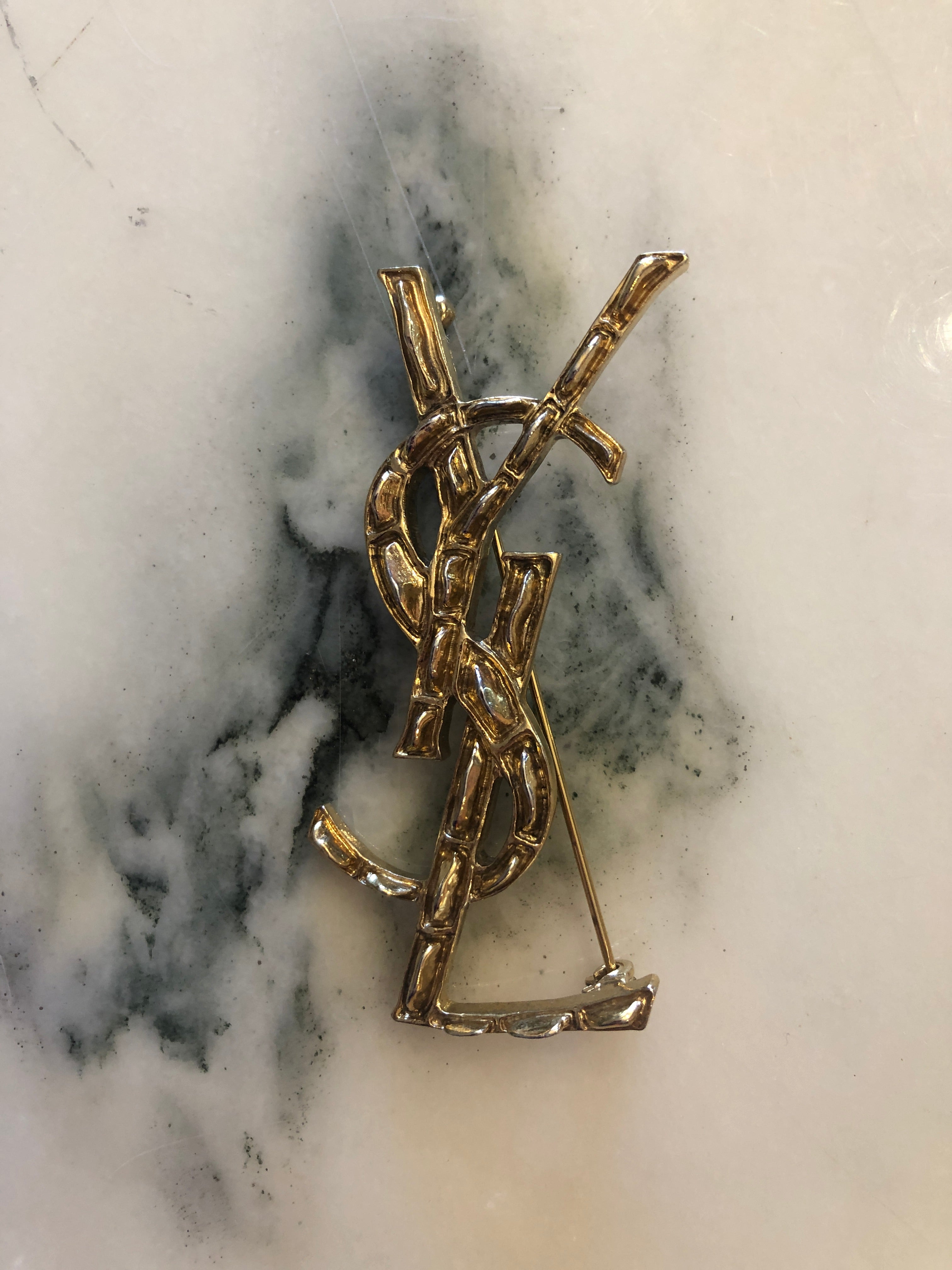 A vintage Yves Saint Laurent gold-tone logo brooch featuring the iconic interlocking YSL monogram in a large sculptural bamboo-effect textured format with polished raised edges, photographed on a white marble surface.