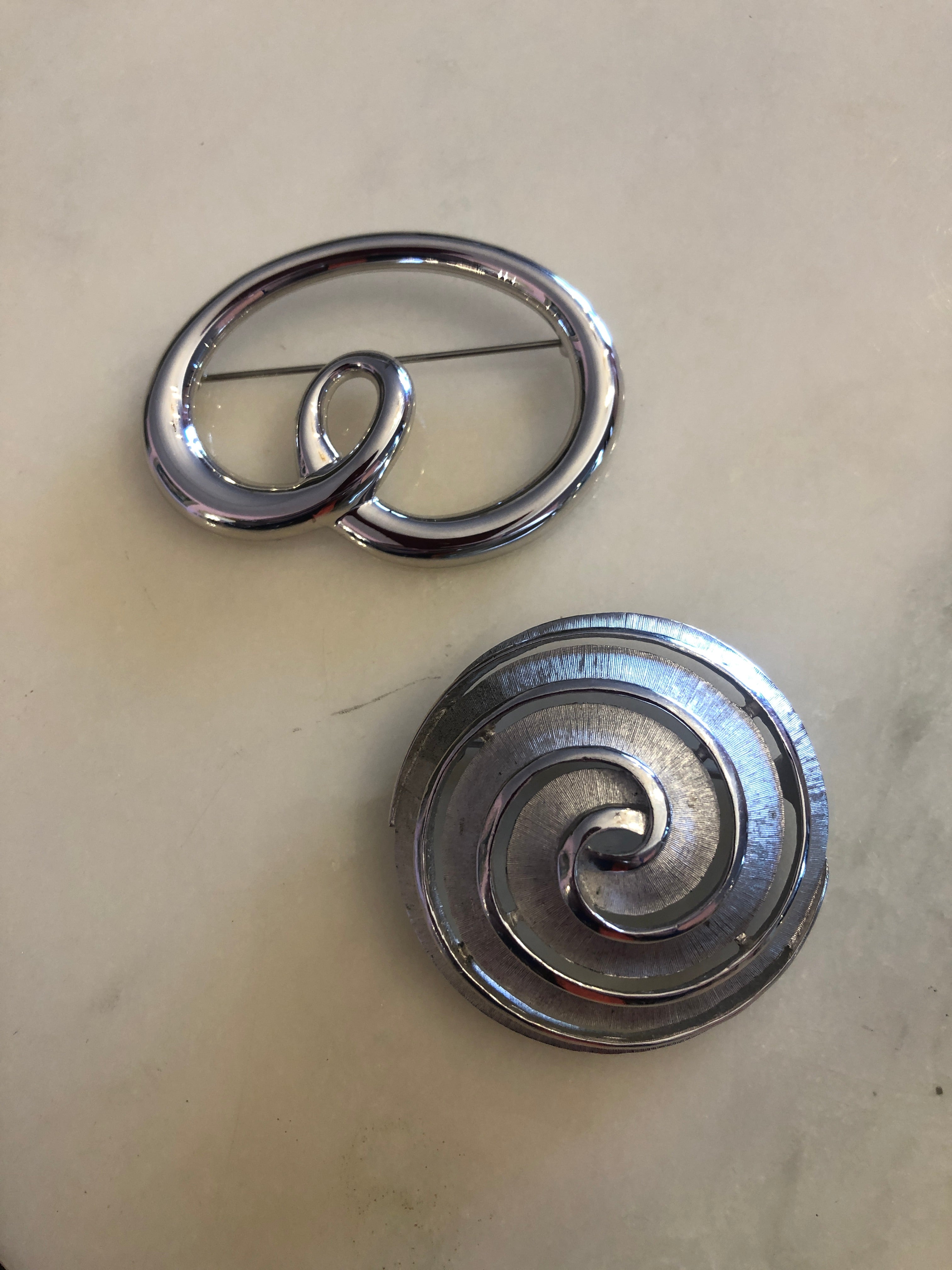 Two vintage silver-tone brooches photographed together on a white marble surface. Top: a Trifari polished silver-tone open loop brooch featuring a fluid cursive swirl form with a small inner loop detail. Bottom: a Monet circular silver-tone brooch with a brushed crosshatch-textured surface and a polished inset spiral motif with a raised outer rim.