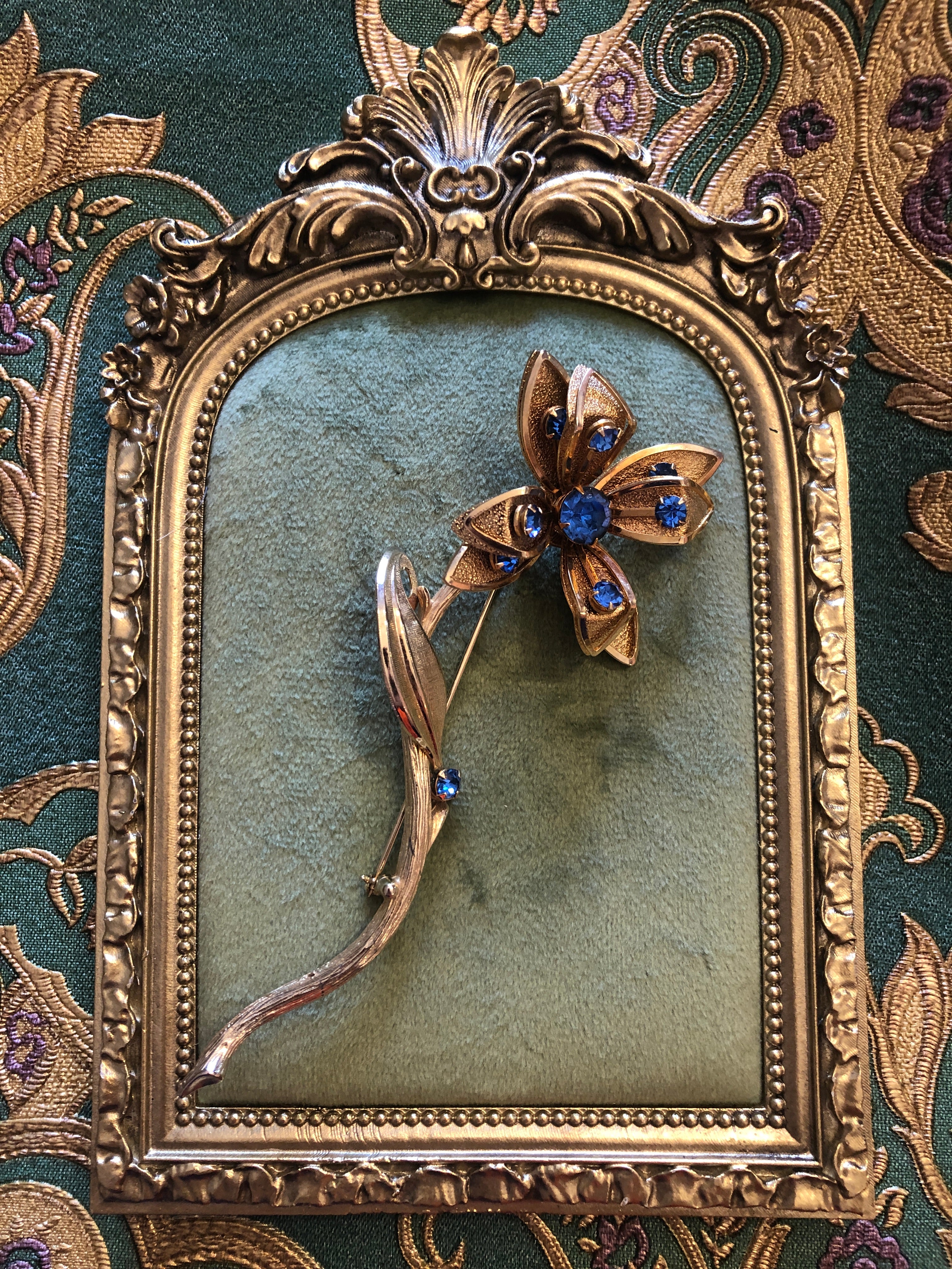 A large vintage gold and silver-tone Fiore Azzurro flower stem brooch featuring five textured gold-tone petals with polished silver-tone tips set with sapphire blue round crystal rhinestones, a central sapphire blue rhinestone cluster, a single silver-tone leaf accent and an additional sapphire blue stone on the elongated curved textured stem, photographed on a green velvet tray with a gold rim. The background is green and gold damask wallpaper