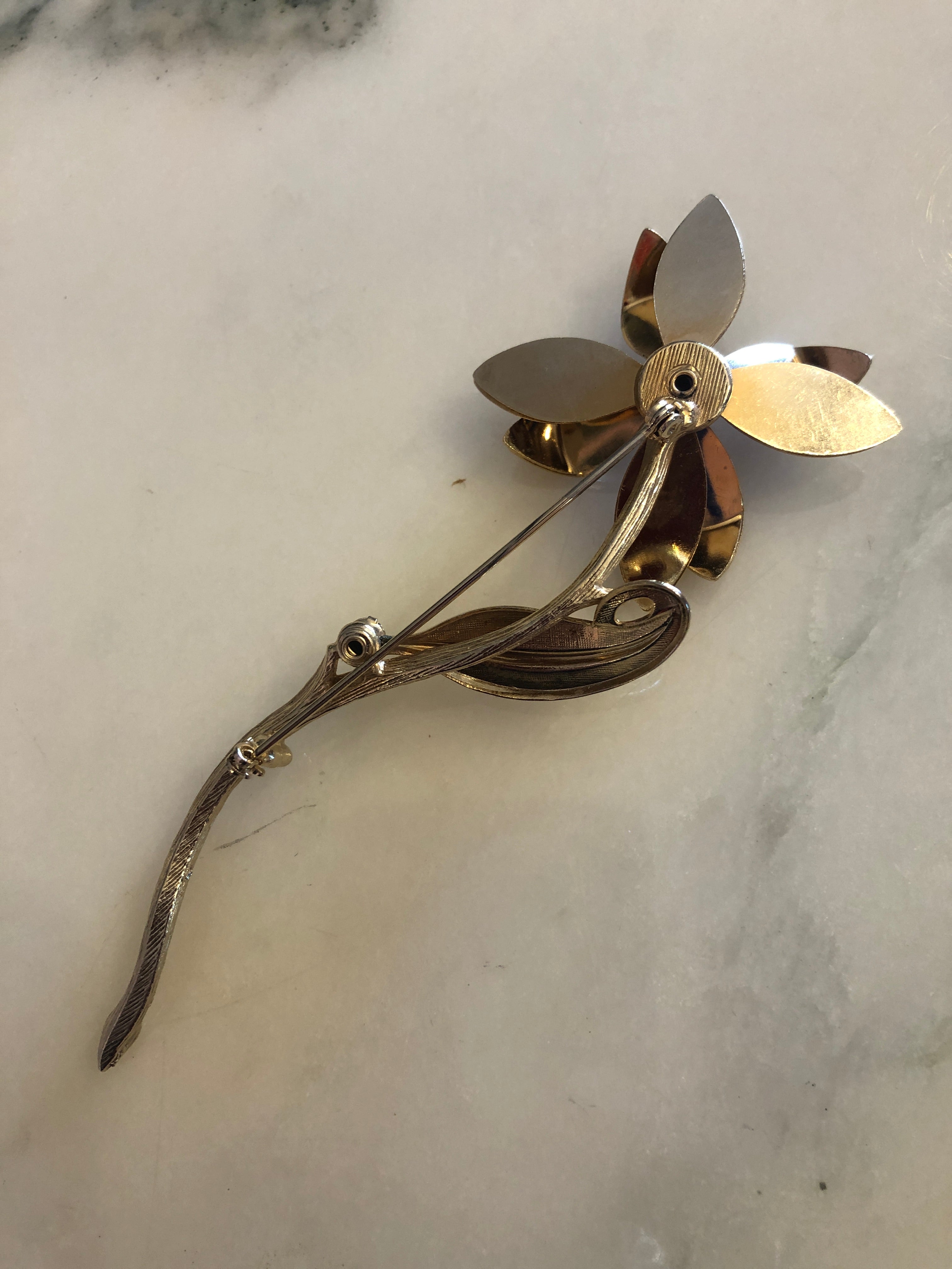 A large vintage gold and silver-tone Fiore Azzurro flower stem brooch featuring five textured gold-tone petals with polished silver-tone tips set with sapphire blue round crystal rhinestones, a central sapphire blue rhinestone cluster, a single silver-tone leaf accent and an additional sapphire blue stone on the elongated curved textured stem, photographed on a white marble surface.
