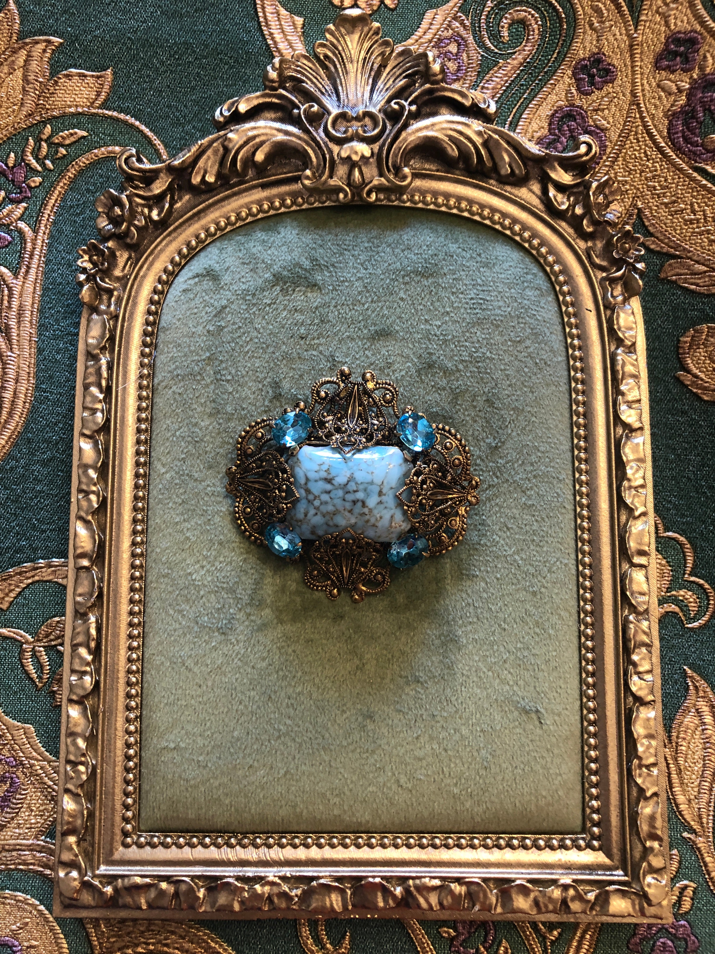 A vintage Czech Azzurra gold-tone filigree brooch featuring a large square turquoise marbled cabochon centre stone set within an ornate granulated and scrollwork openwork frame, accented with six faceted aqua blue crystal rhinestones at the cardinal points, photographed on a green velvet tray with a gold frame.  The background is green and gold damask wallpaper