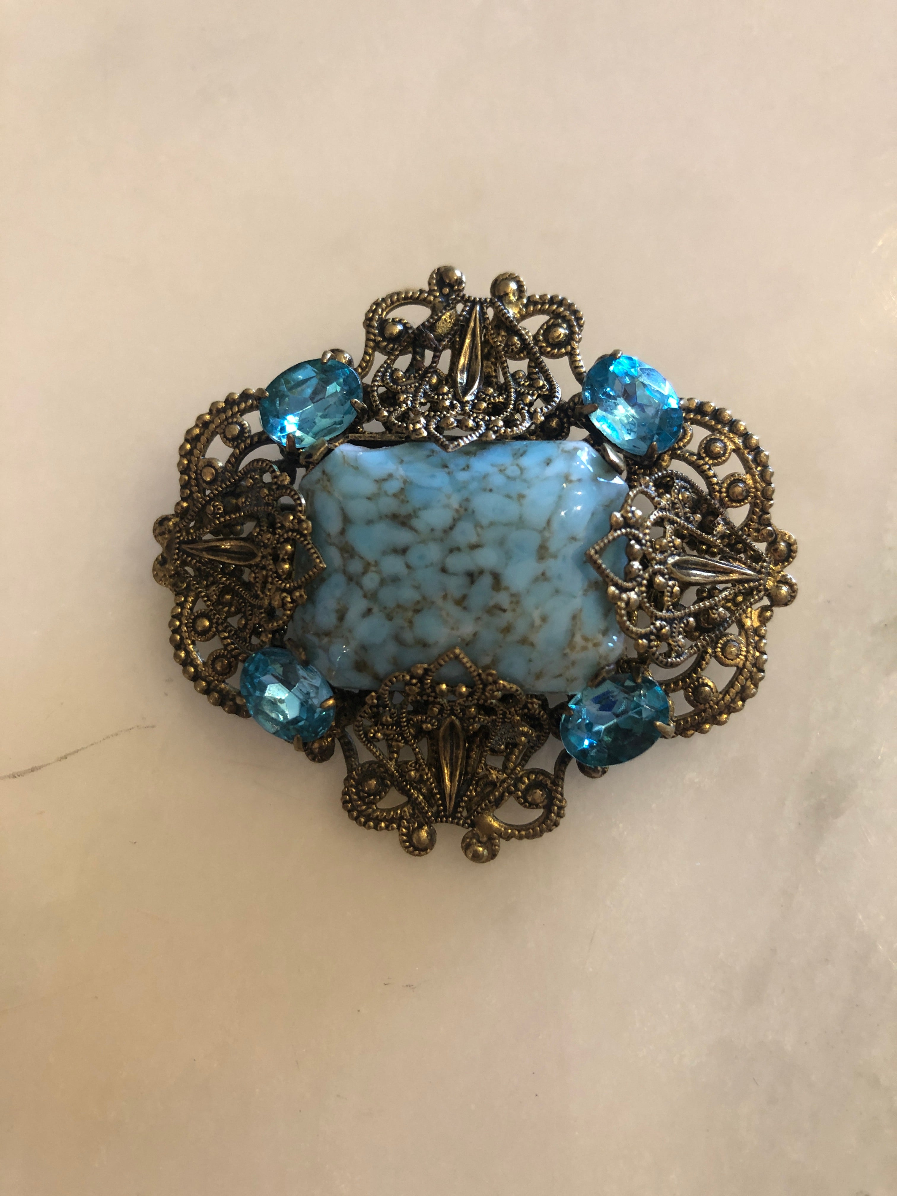A vintage Czech Azzurra gold-tone filigree brooch featuring a large square turquoise marbled cabochon centre stone set within an ornate granulated and scrollwork openwork frame, accented with six faceted aqua blue crystal rhinestones at the cardinal points, photographed on a white marble surface.