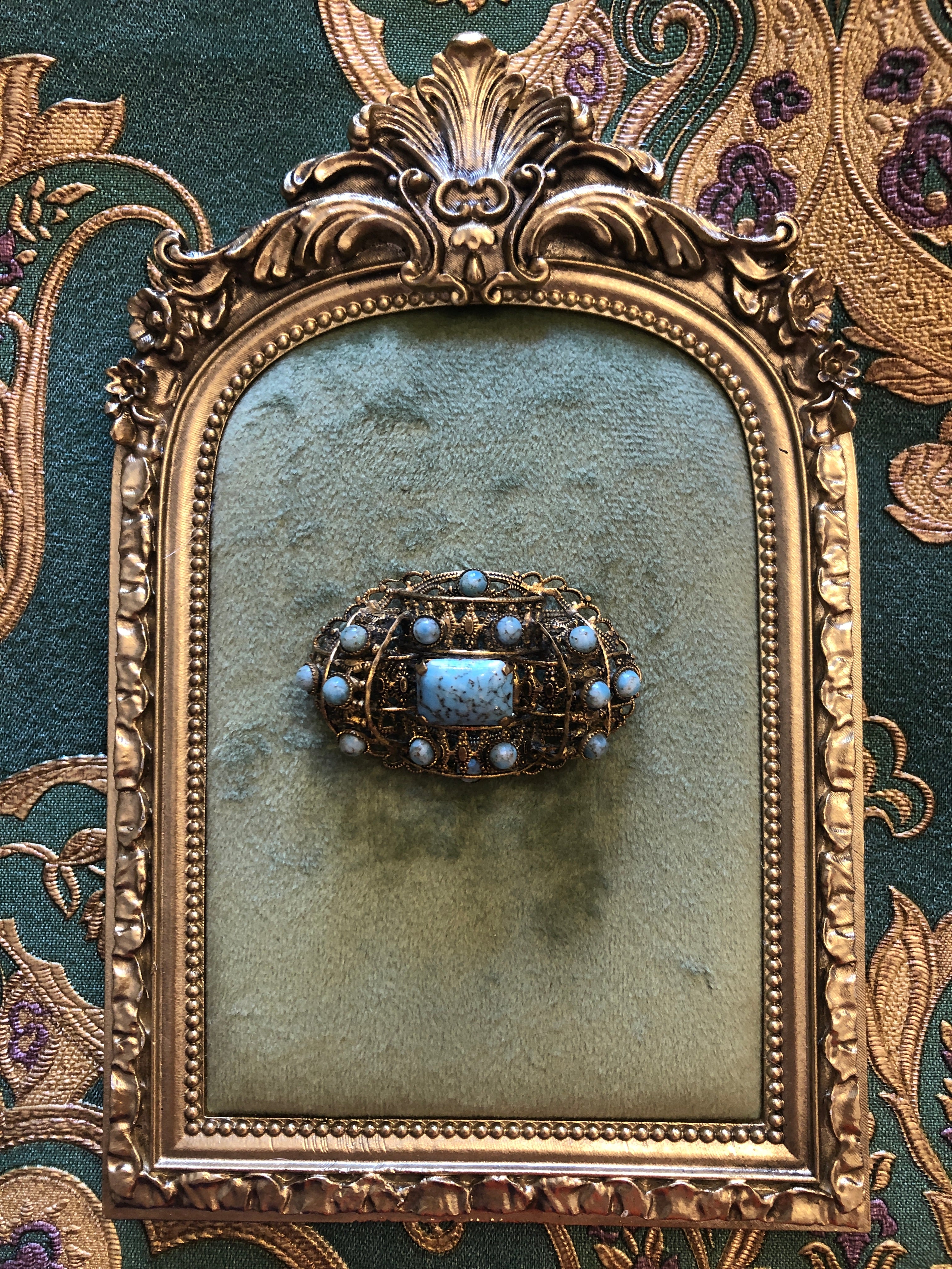 A vintage Czech Tesoro Azzurro gold-tone oval filigree brooch featuring a square turquoise marbled cabochon centre stone set within an intricate openwork granulated scrollwork frame, surrounded by fourteen round turquoise marbled cabochon stones set along the outer border, photographed on a white marble surface.