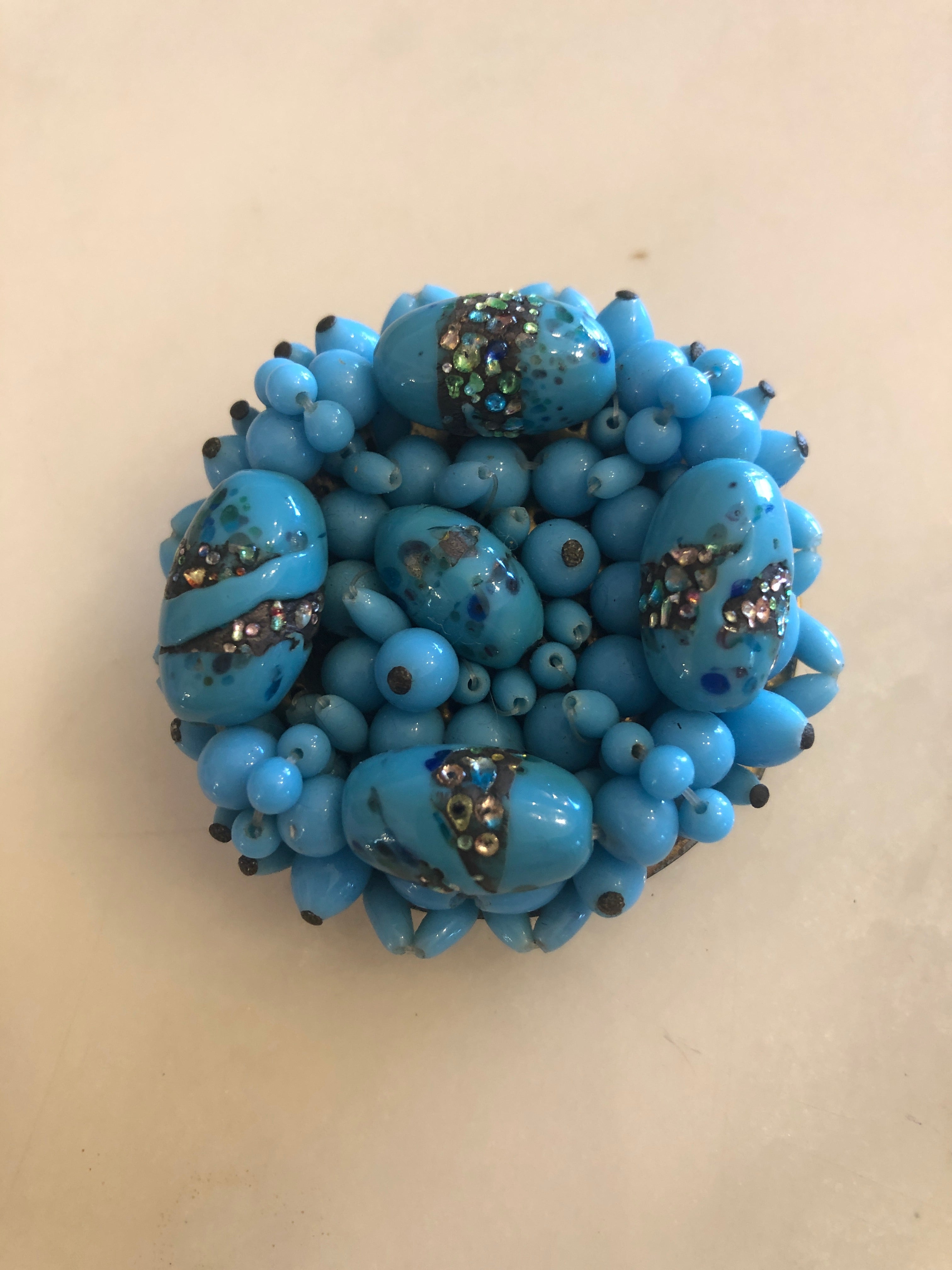 A vintage Laguna Turchese circular cluster brooch featuring a dense domed arrangement of graduating powder blue glass beads in various sizes, interspersed with four larger oval focal beads decorated with aurora borealis crystal rhinestone and metallic foil encrusted band details, photographed on a white marble surface.