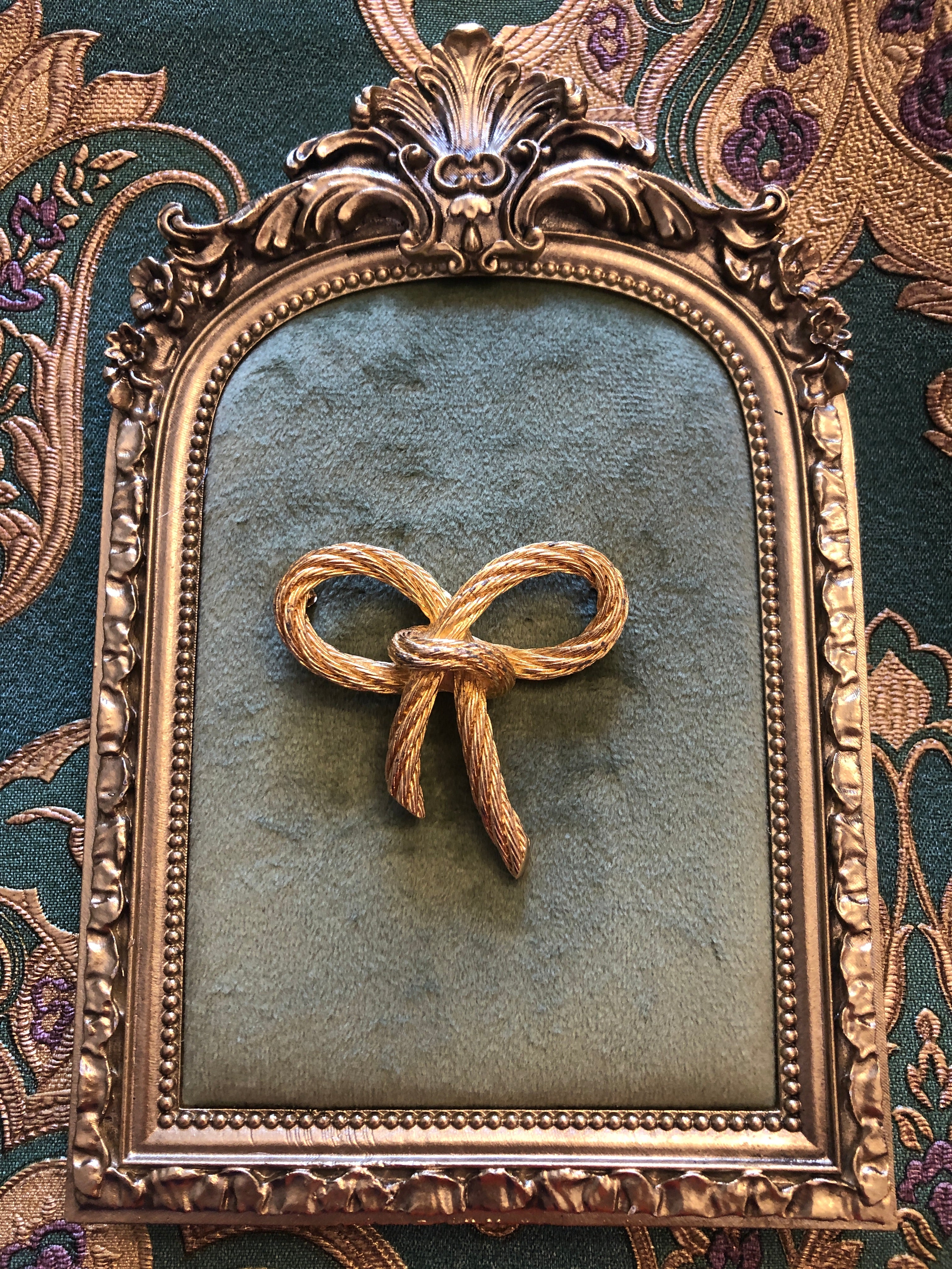 A vintage Christian Dior gold-tone bow brooch crafted from finely detailed twisted rope texture metal forming two open looped wings tied at the centre with trailing double tails, photographed on a green velvet tray with a gold frame. The background is green and gold damask wallpaper