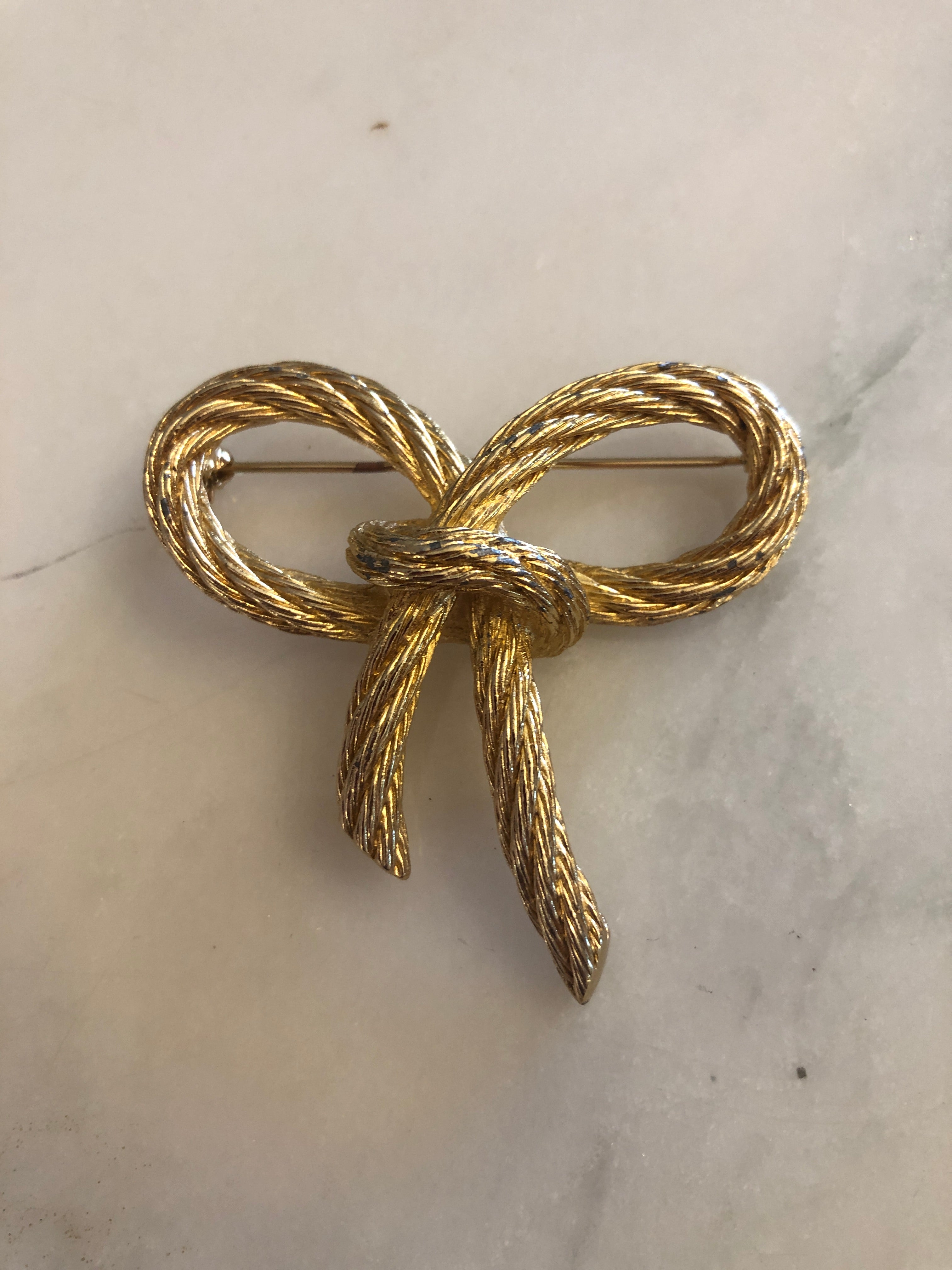 A vintage Christian Dior gold-tone bow brooch crafted from finely detailed twisted rope texture metal forming two open looped wings tied at the centre with trailing double tails, photographed on a white marble surface.