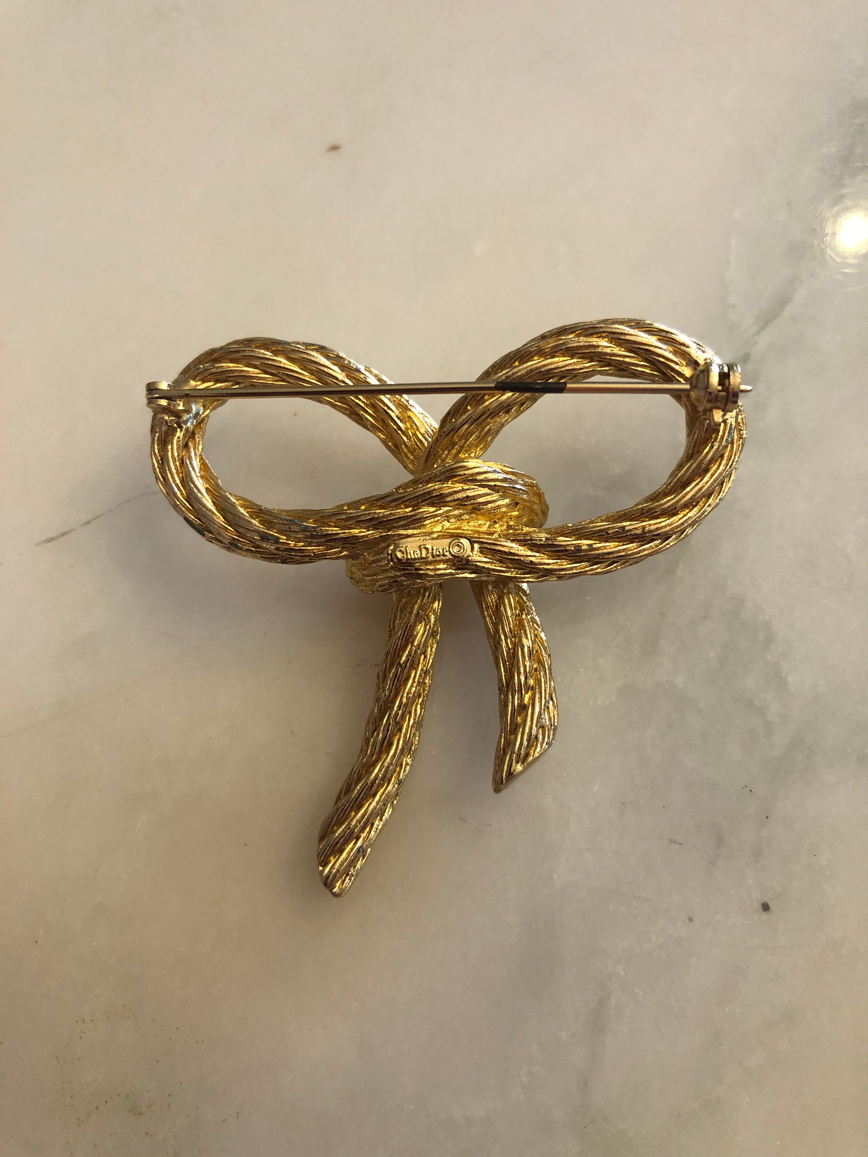 A vintage Christian Dior gold-tone bow brooch crafted from finely detailed twisted rope texture metal forming two open looped wings tied at the centre with trailing double tails, photographed on a white marble surface.