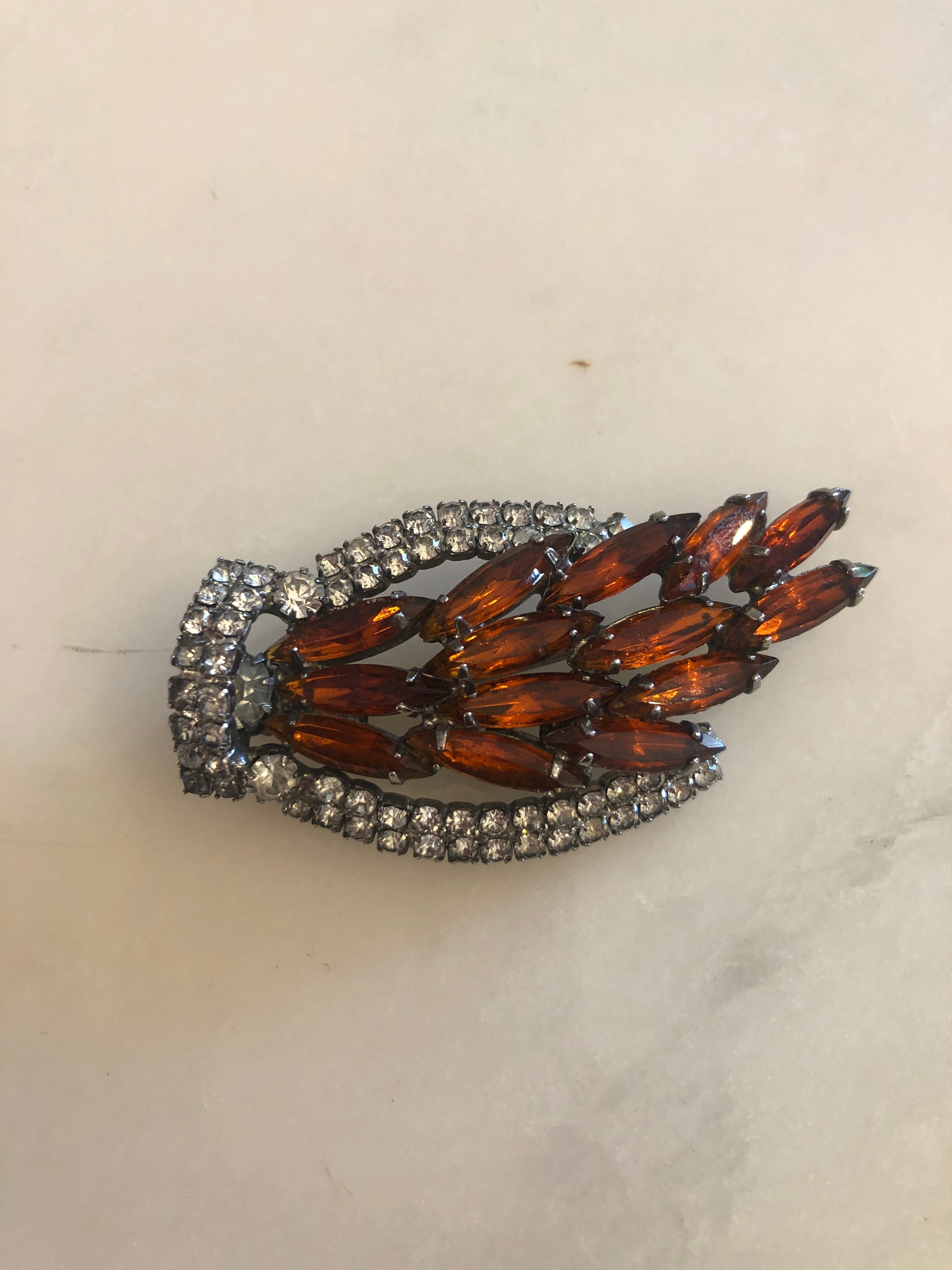 A 3cm x 6cm vintage dark silver-tone flame-shaped brooch with layered deep amber navette-cut crystals bordered by a curved double row of clear round crystal rhinestones, photographed on a white marble surface.