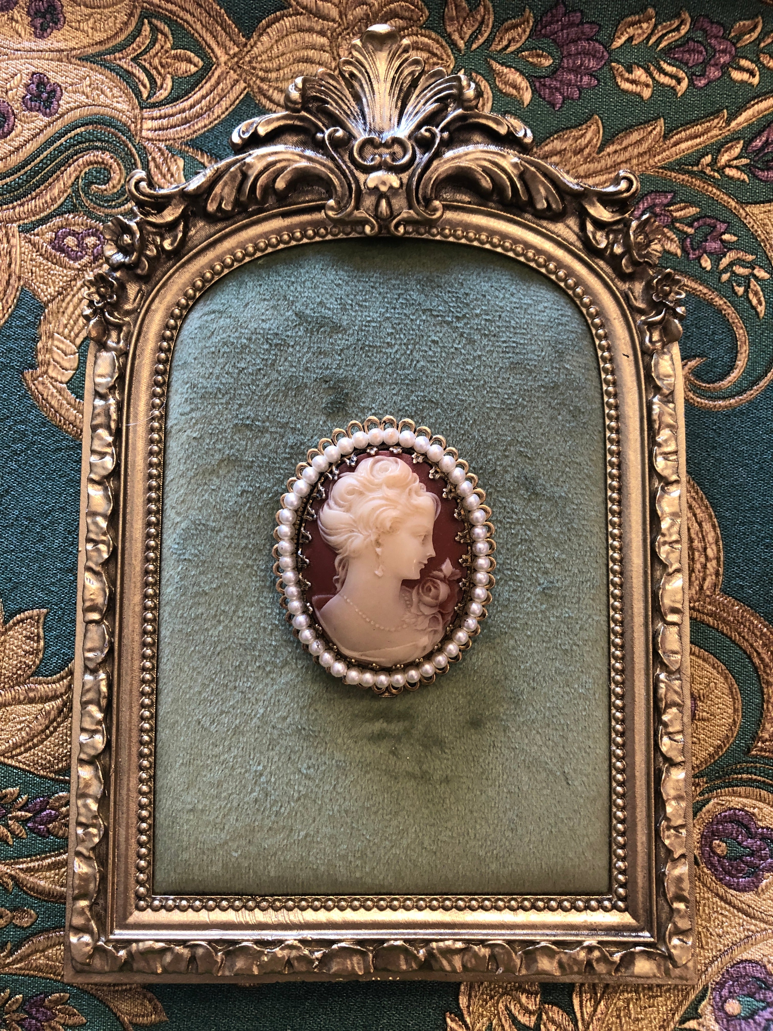 A 5.5cm x 4cm vintage cameo brooch with a carved cream portrait of a lady with upswept curls, drop earring and rose detail on a blush pink ground, set in a gold-tone filigree frame with a full seed pearl border. Photographed on a green velvet tray, with an ornate gold frame. The background is green and gold damask wallpaper. 