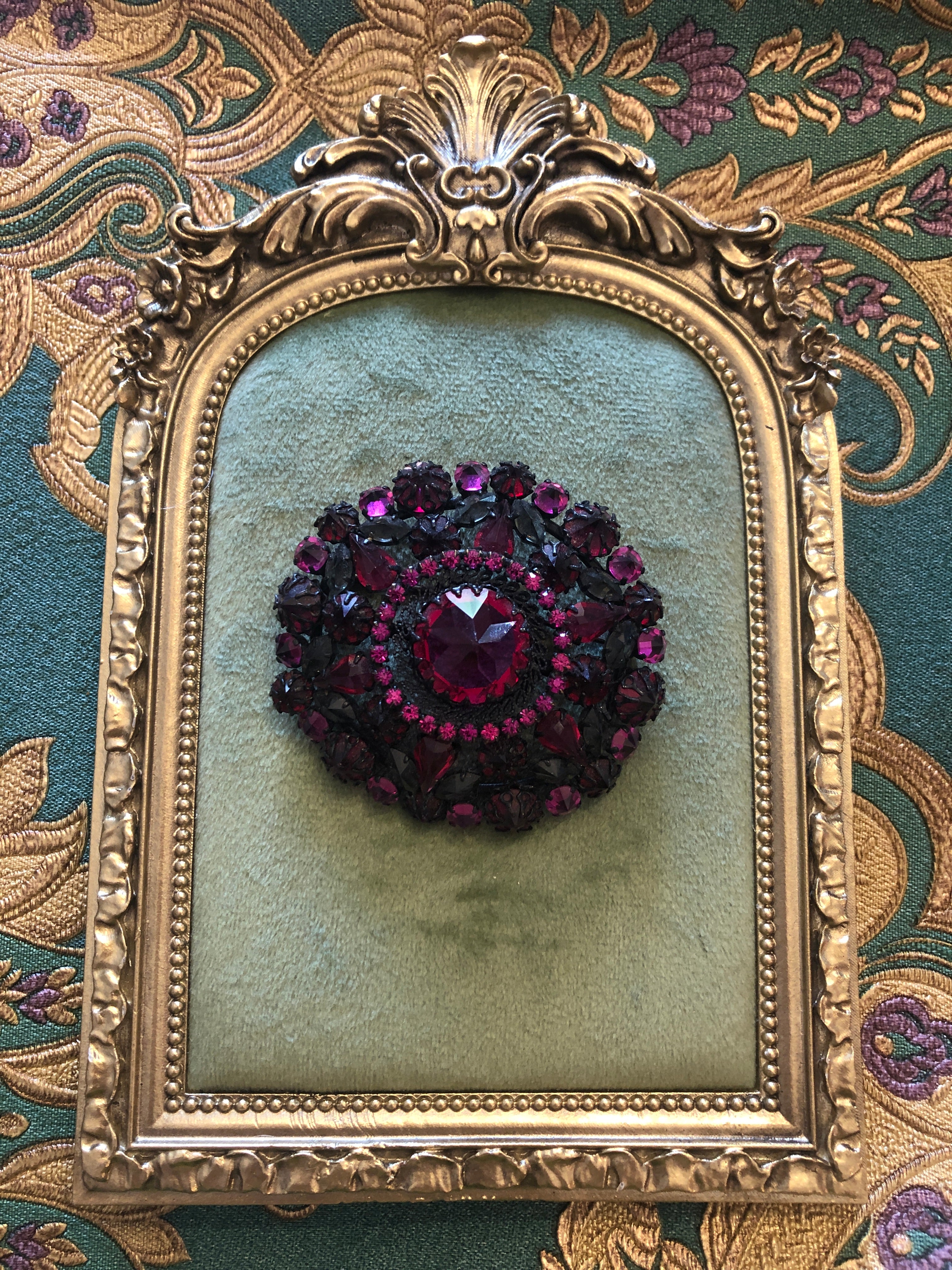 A 6cm x 6cm vintage oxidised black-tone circular brooch with a large faceted ruby red oval centre stone surrounded by layered rings of hot magenta and purple pear-cut and round crystal rhinestones. Photographed on a green velvet tray, with an ornate gold frame. The background is green and gold damask wallpaper