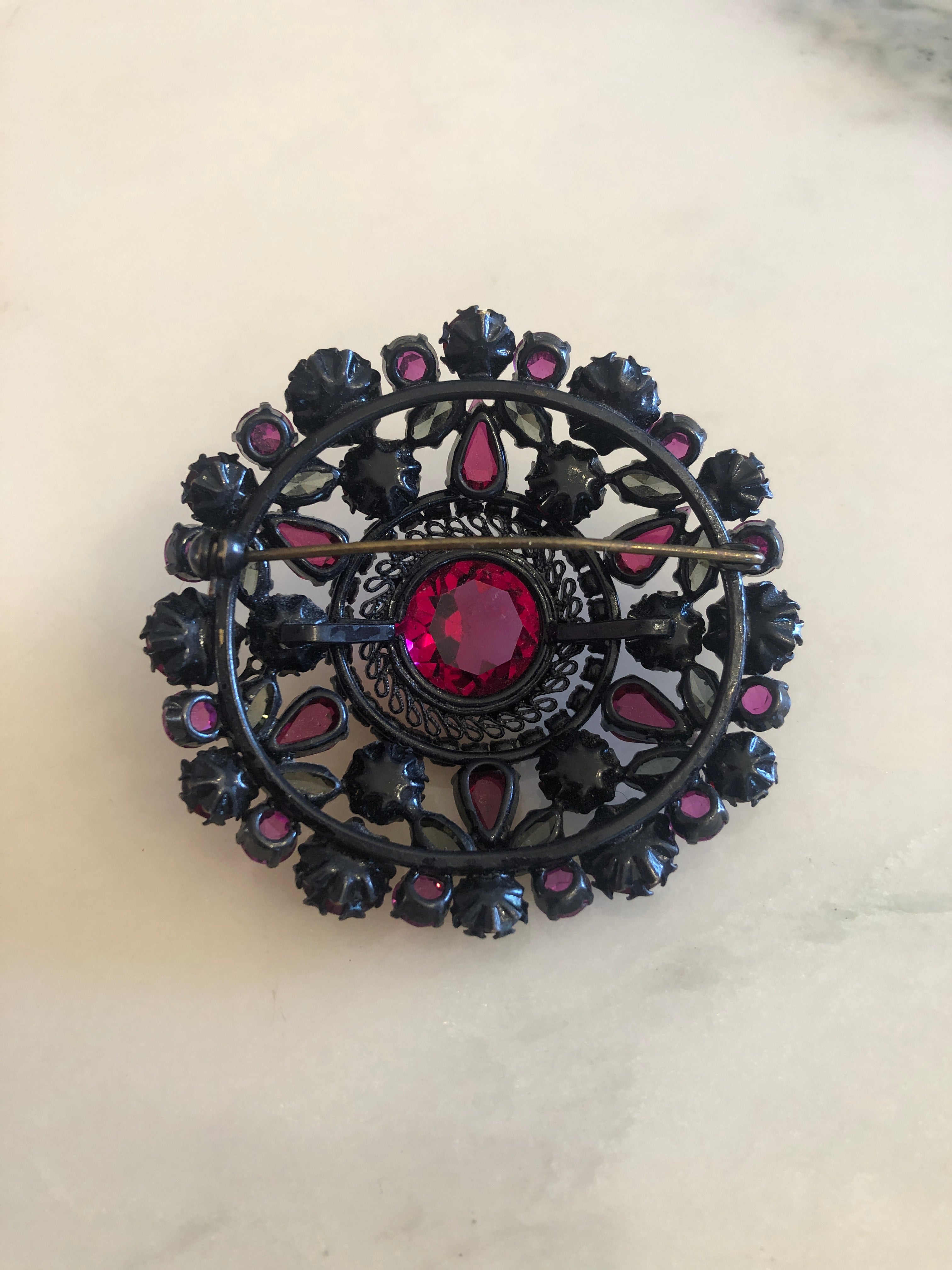 A 6cm x 6cm vintage oxidised black-tone circular brooch with a large faceted ruby red oval centre stone surrounded by layered rings of hot magenta and purple pear-cut and round crystal rhinestones, photographed on a white marble surface.