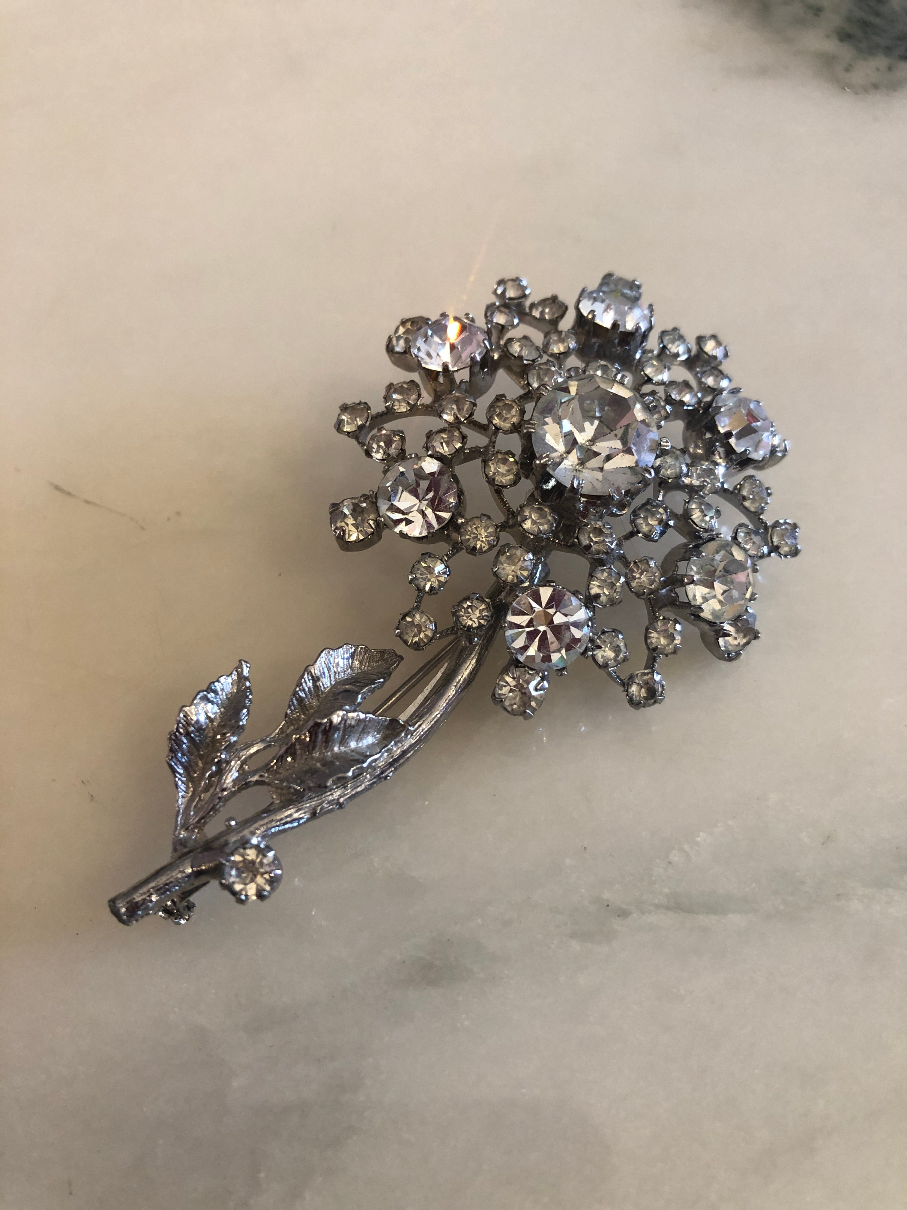 A 7.5cm x 4.5cm vintage silver-tone crystal floral spray brooch with a graduating clear round crystal rhinestone flower head on fine wire stems and a textured silver-tone leaf and stem base, photographed on a white marble surface.