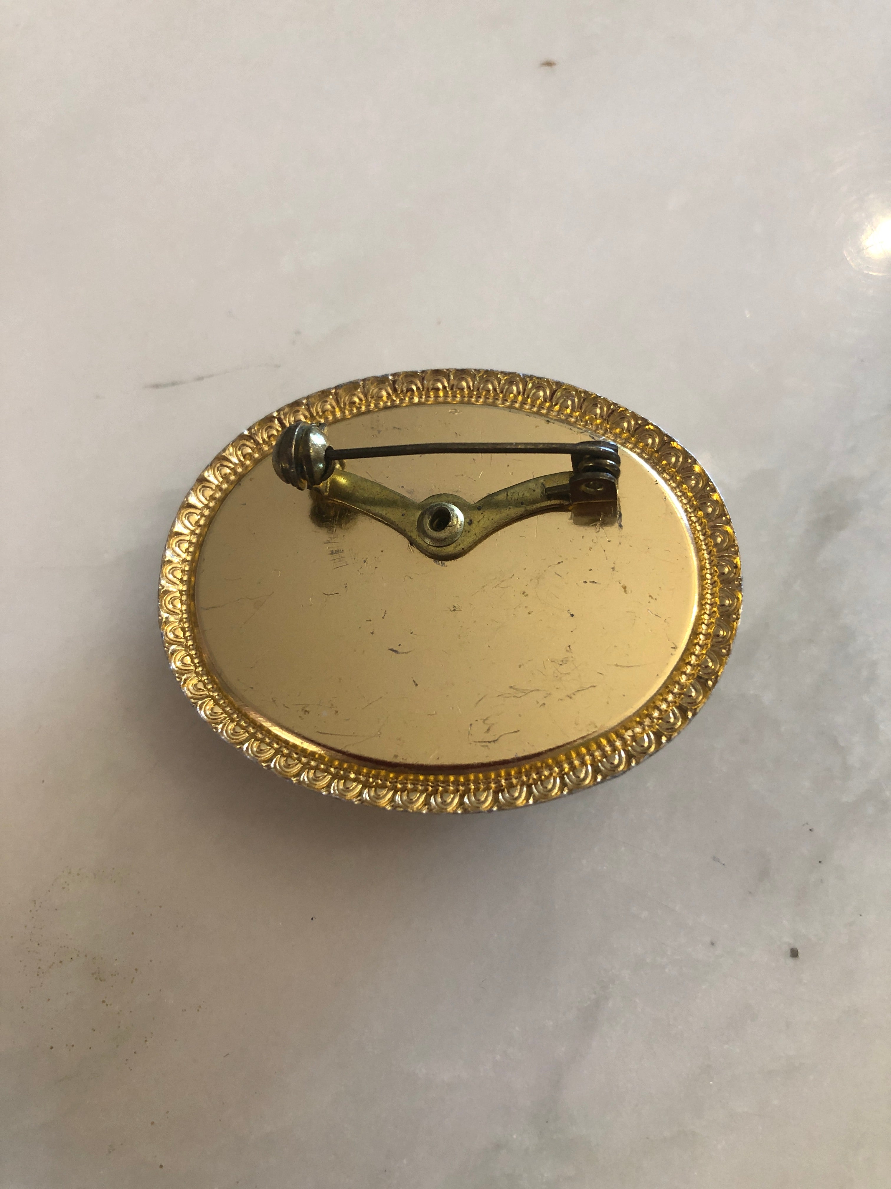 A 3.5cm x 4.5cm vintage cameo brooch with a carved white classical portrait on a deep blue ground, with flowing hair and flower bud detail, set in a gold-tone scrollwork frame, photographed on a white marble surface.