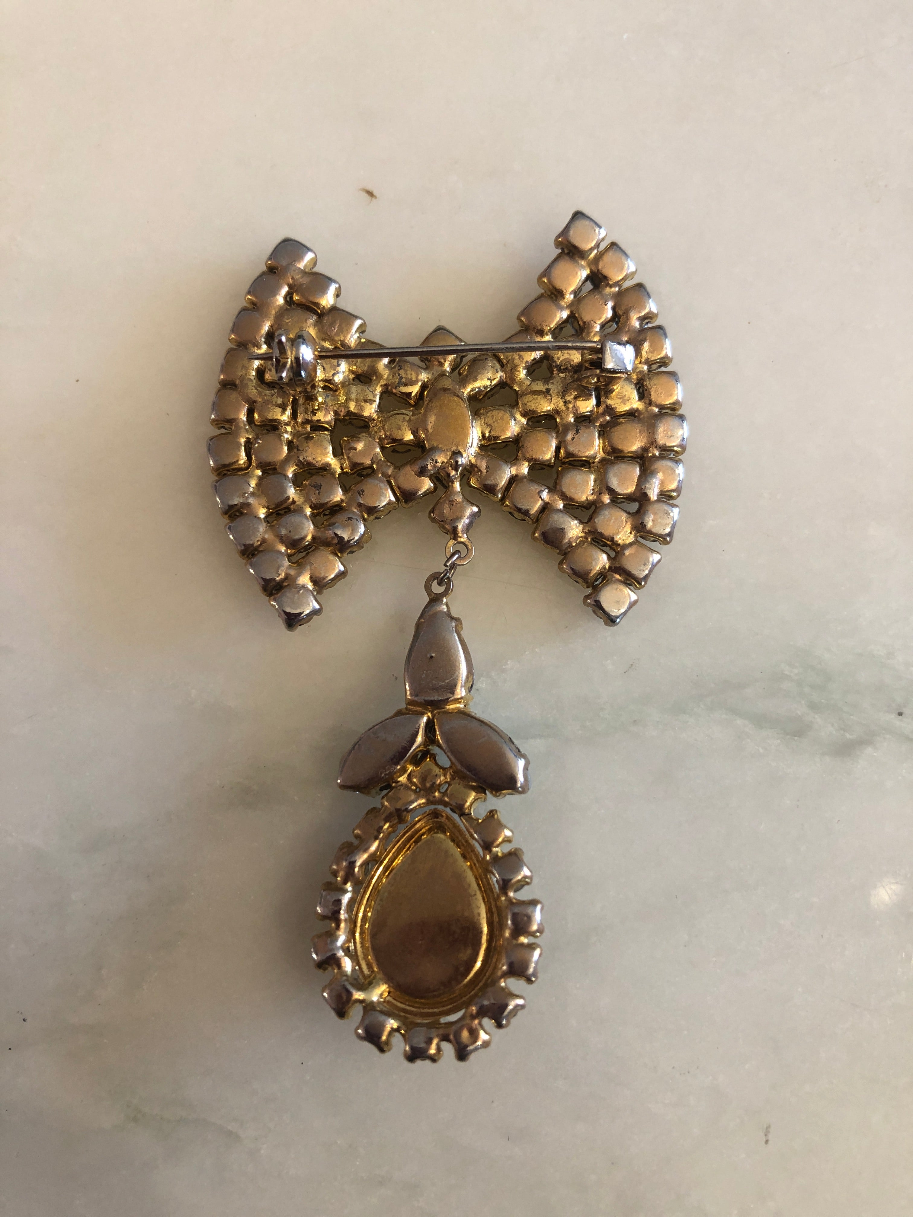 A 6.5cm x 4cm vintage gold-tone bow brooch densely pavé-set with clear crystal rhinestones, with a suspended drop of clear pear and marquise-cut crystals leading to a silver-tone teardrop cabochon, photographed on a white marble surface.