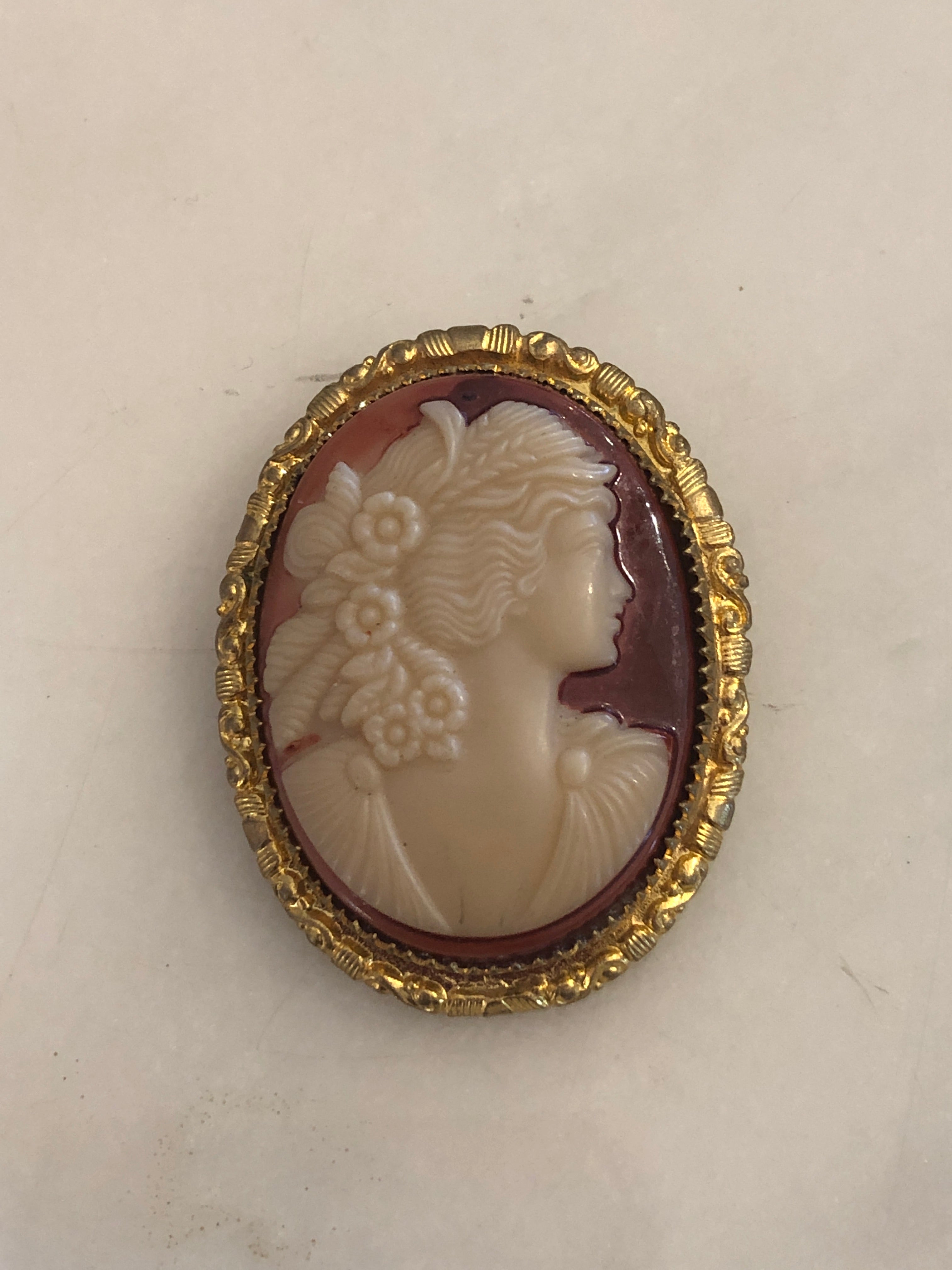 A 4cm x 3cm vintage shell cameo brooch with a carved cream relief portrait of a flower-crowned goddess in profile on a pink-mauve ground, set in a gold-tone scrollwork frame, photographed on a white surface.