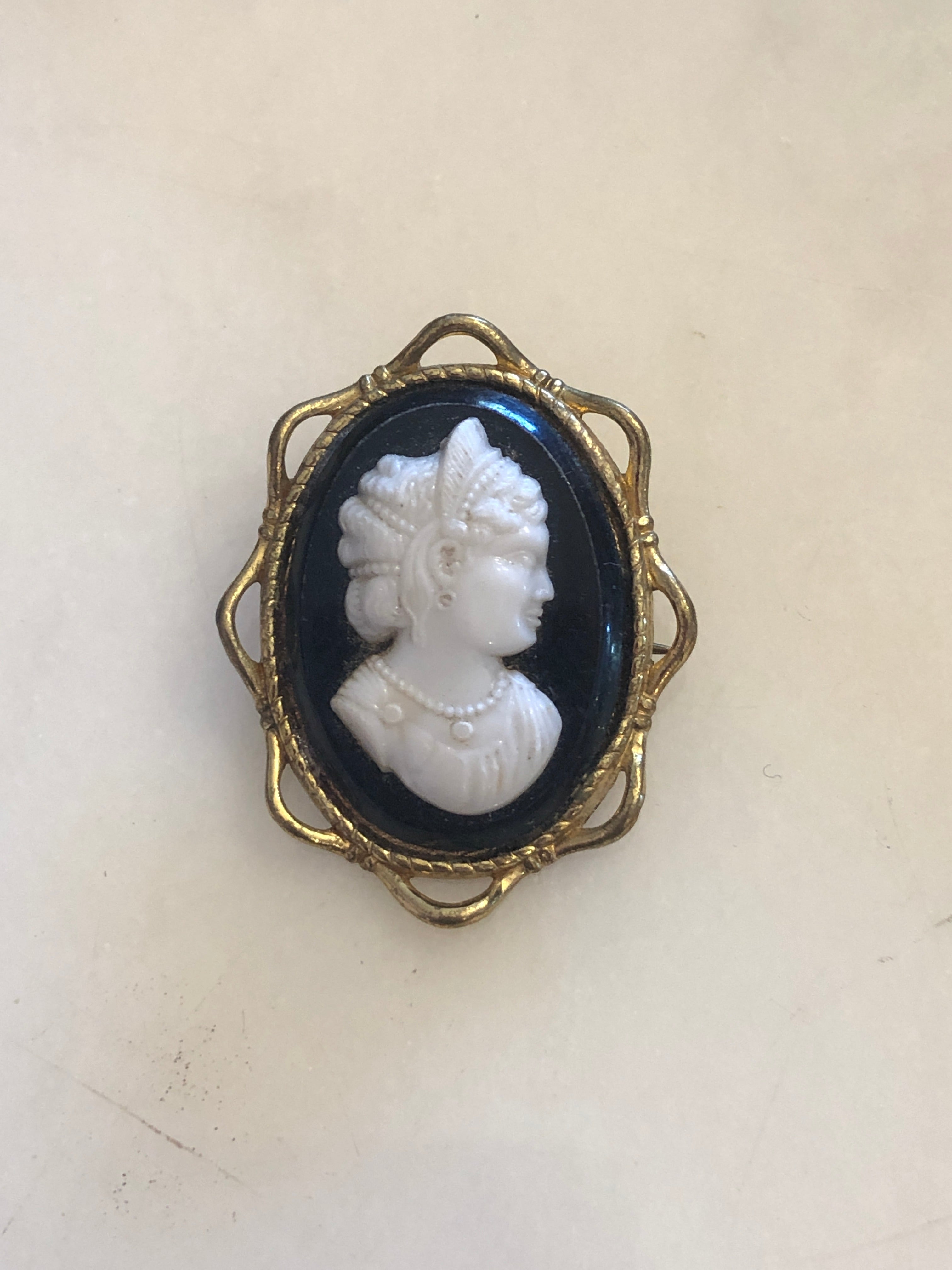 A 4cm x 3cm vintage black and white cameo brooch featuring a carved white relief classical portrait of a noblewoman in profile with elaborate feathered and pearl upswept hair and a pearl necklace, set on a deep black oval ground within a gold-tone openwork filigree frame, photographed on a white marble surface.