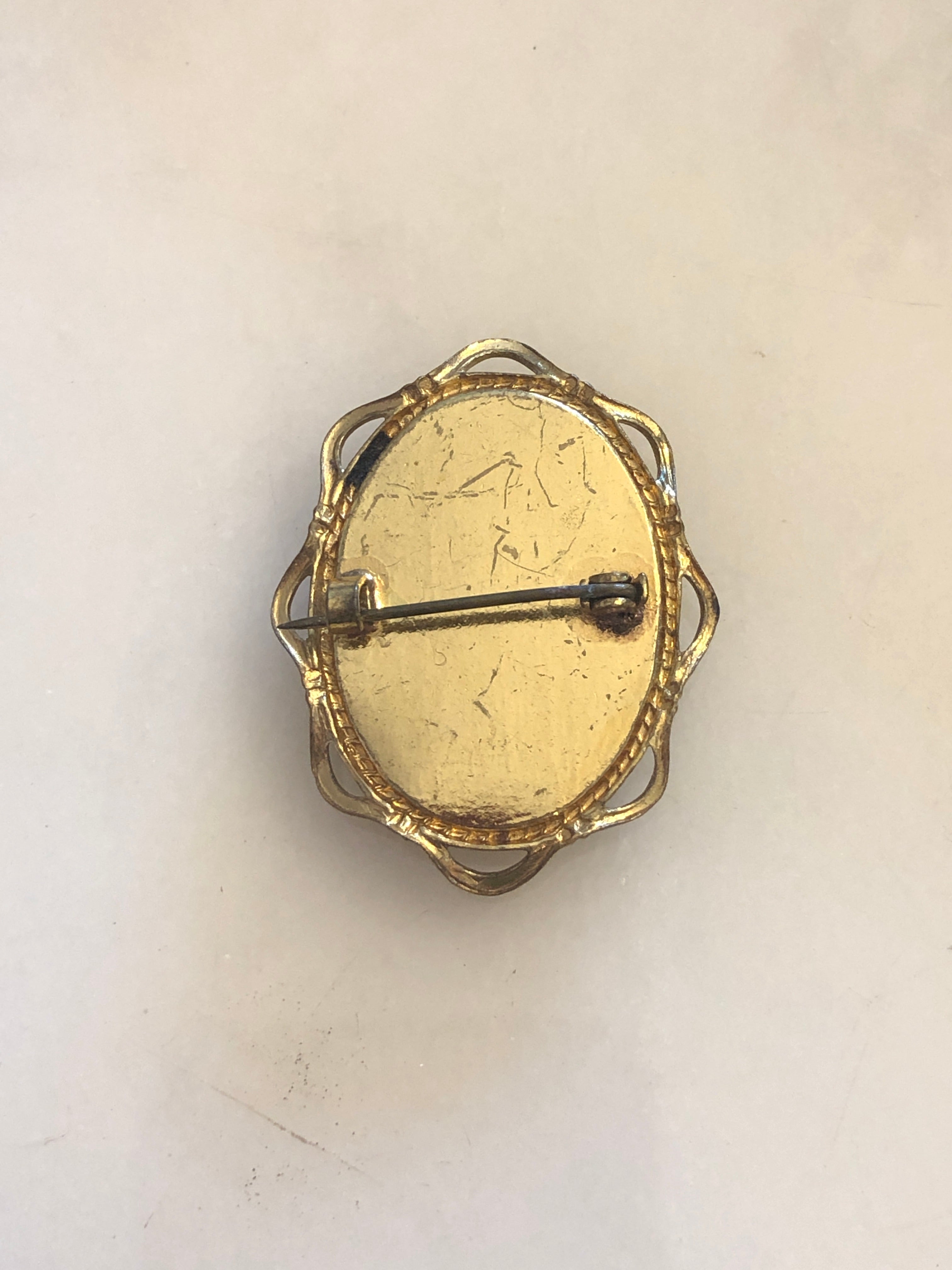 A 4cm x 3cm vintage black and white cameo brooch featuring a carved white relief classical portrait of a noblewoman in profile with elaborate feathered and pearl upswept hair and a pearl necklace, set on a deep black oval ground within a gold-tone openwork filigree frame, photographed on a white marble surface.