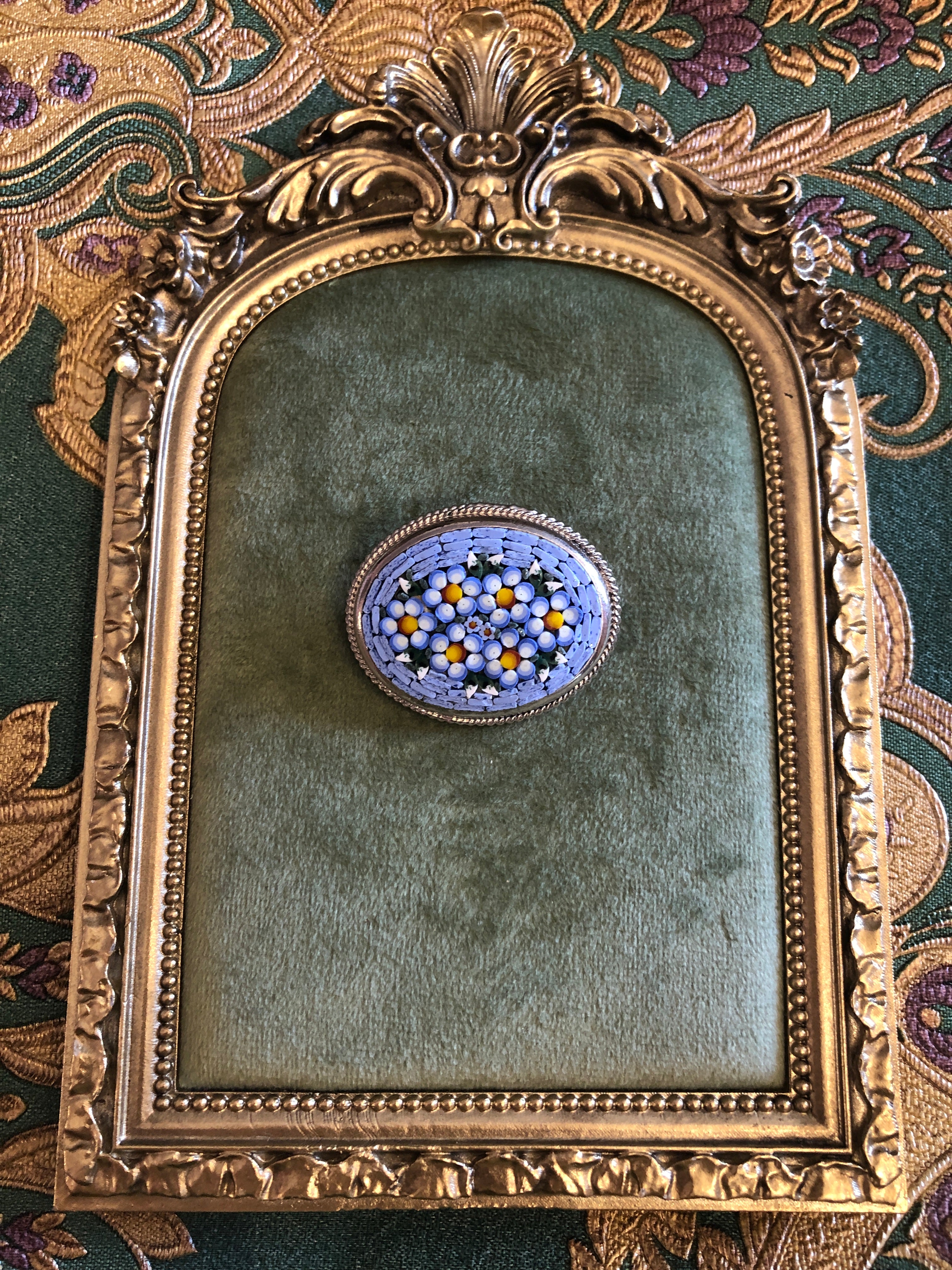 A 3cm x 4cm vintage Italian micro mosaic oval brooch featuring a hand assembled glass tessera design of forget-me-not flowers in cornflower blue, white and yellow on a blue ground, set within a silver-tone frame with twisted rope edge detail, photographed on a green velvet tray, with a gold frame. The background is green and gold damask wallpaper.