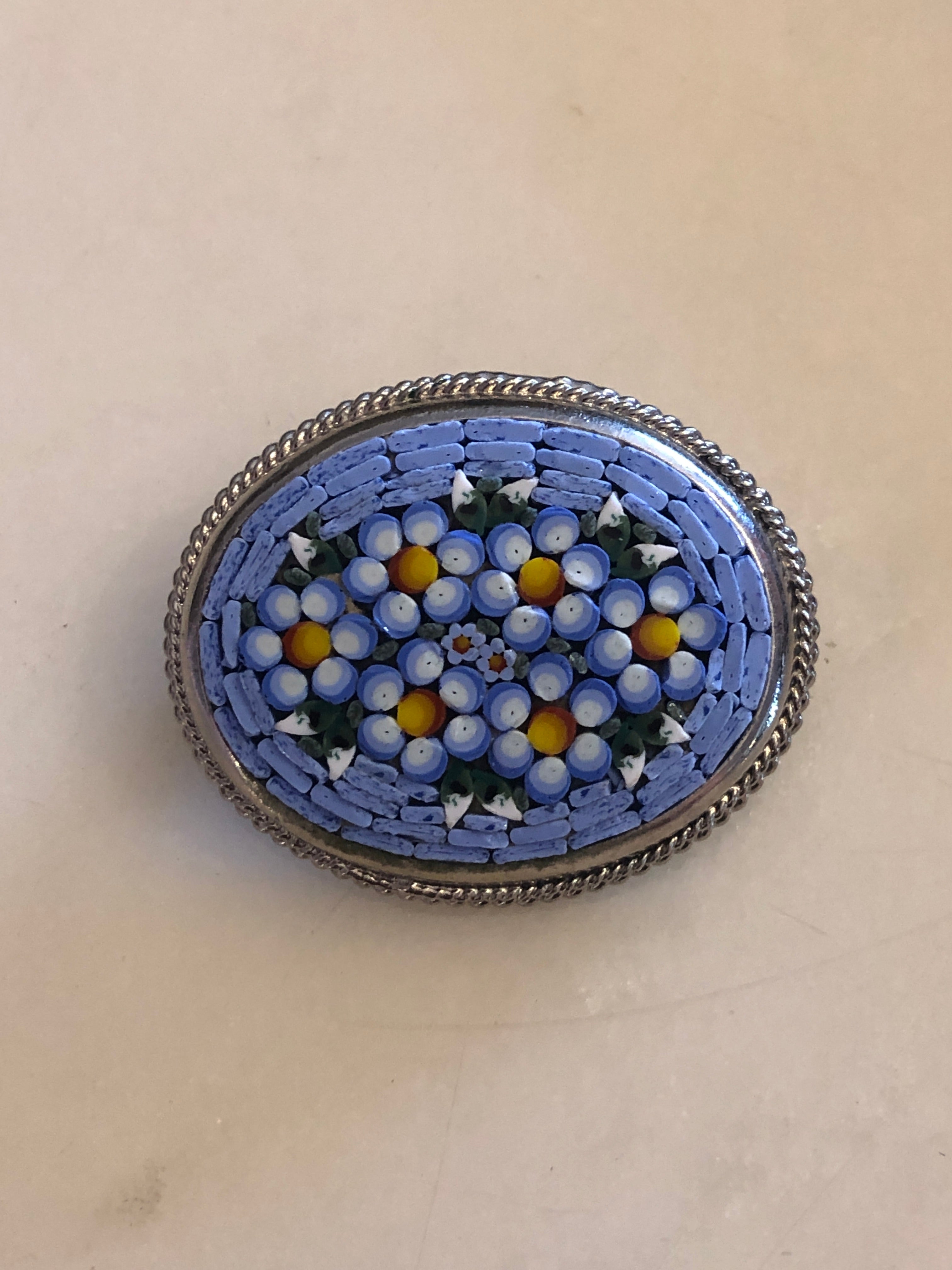 A 3cm x 4cm vintage Italian micro mosaic oval brooch featuring a hand assembled glass tessera design of forget-me-not flowers in cornflower blue, white and yellow on a blue ground, set within a silver-tone frame with twisted rope edge detail, photographed on a white marble surface.