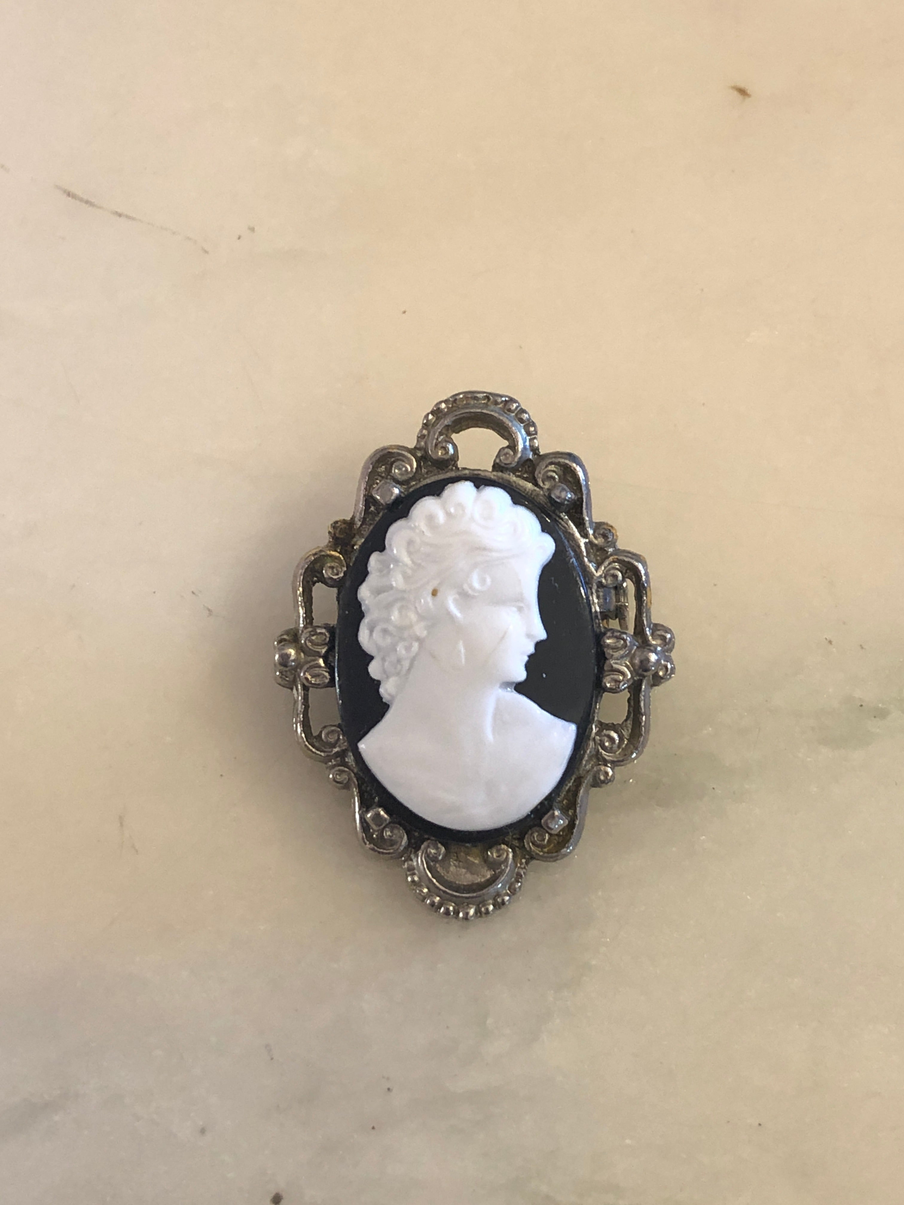 A 3.5cm x 2.5cm vintage black and white cameo brooch with a carved white classical portrait on a black oval ground, set in a silver-tone scrollwork frame, photographed on a white marble surface.