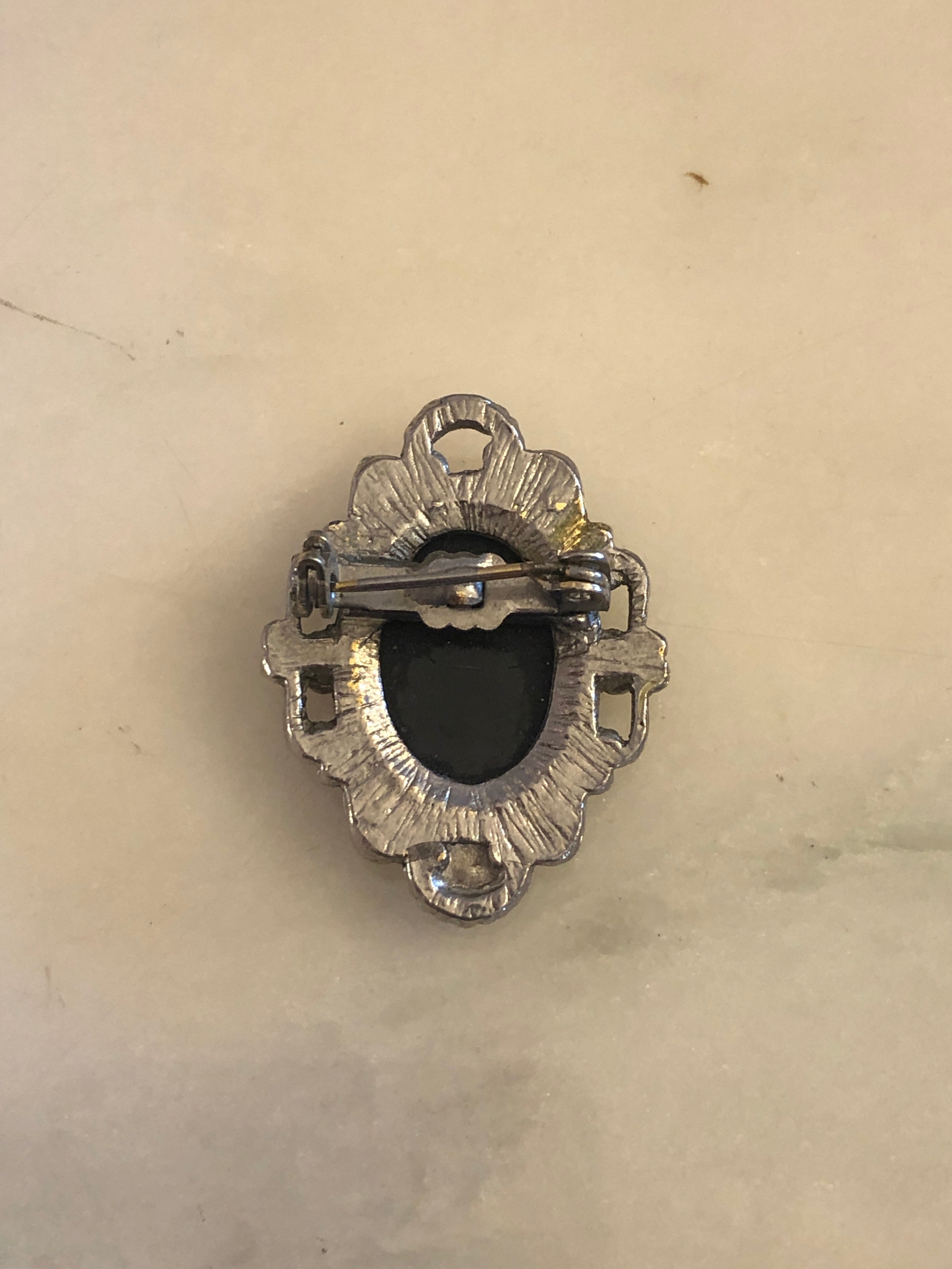 A 3.5cm x 2.5cm vintage black and white cameo brooch with a carved white classical portrait on a black oval ground, set in a silver-tone scrollwork frame, photographed on a white marble surface.