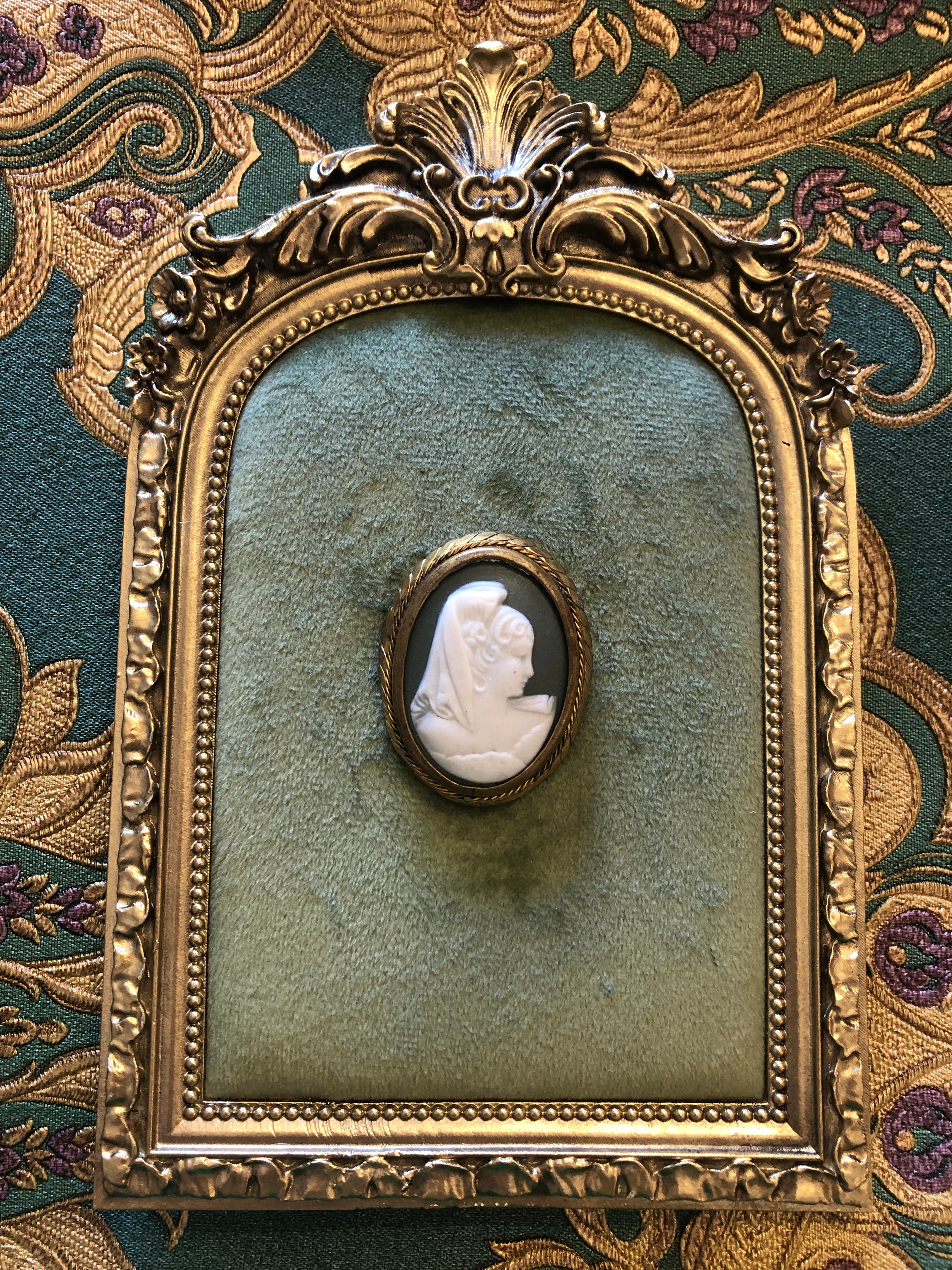 A 4cm x 3.5cm vintage cameo brooch with a carved white veiled classical portrait on a deep green ground in a gold-tone twisted rope frame, photographed on a white marble surface.