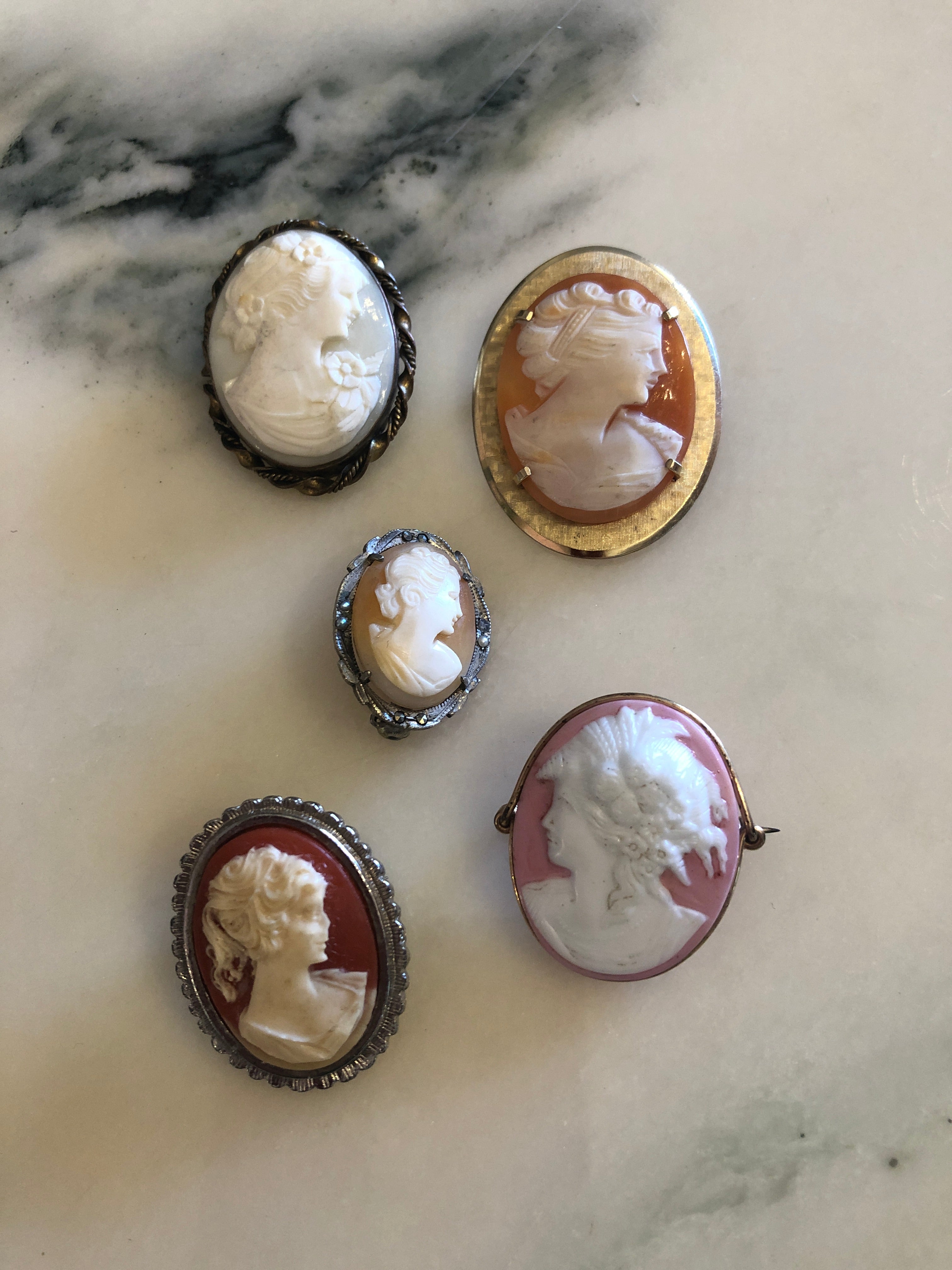 Five vintage cameo brooches displayed on sage green velvet, each with a carved classical lady portrait on grounds of white, blush, peach, terracotta and rose pink in mixed silver and gold-tone frames.
