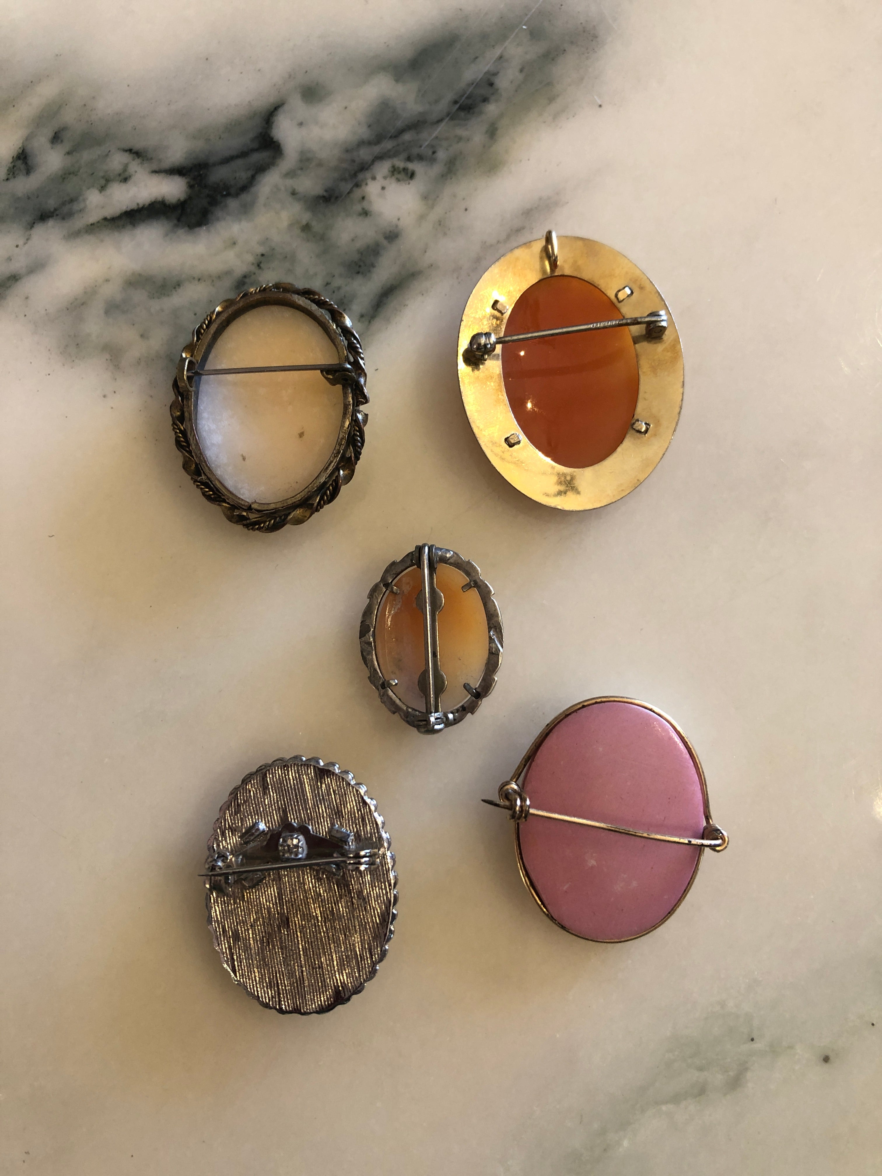 Five vintage cameo brooches displayed on sage green velvet, each with a carved classical lady portrait on grounds of white, blush, peach, terracotta and rose pink in mixed silver and gold-tone frames.
