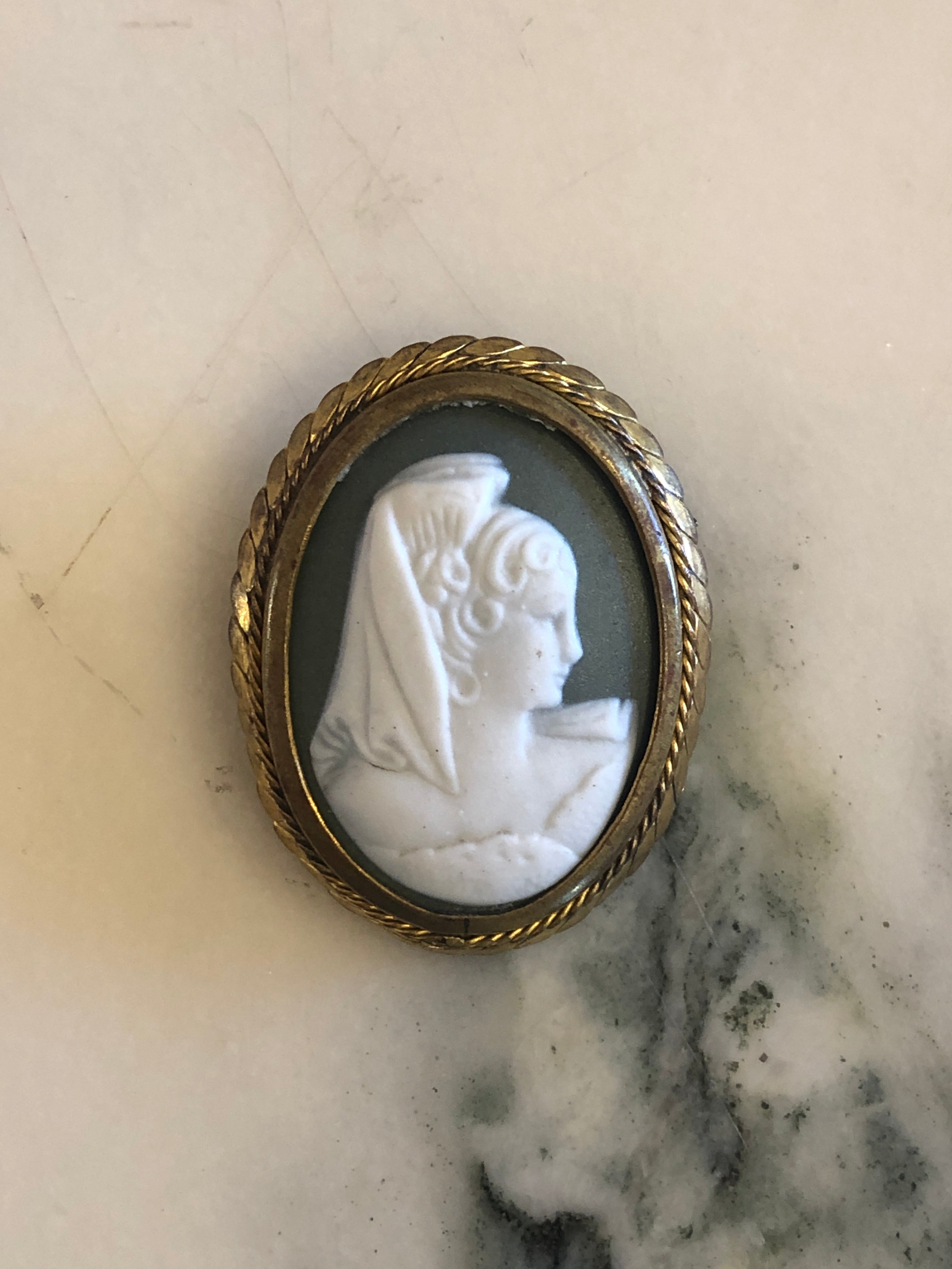 A 4cm x 3.5cm vintage cameo brooch with a carved white veiled classical portrait on a deep green ground in a gold-tone twisted rope frame, photographed on a white marble surface.