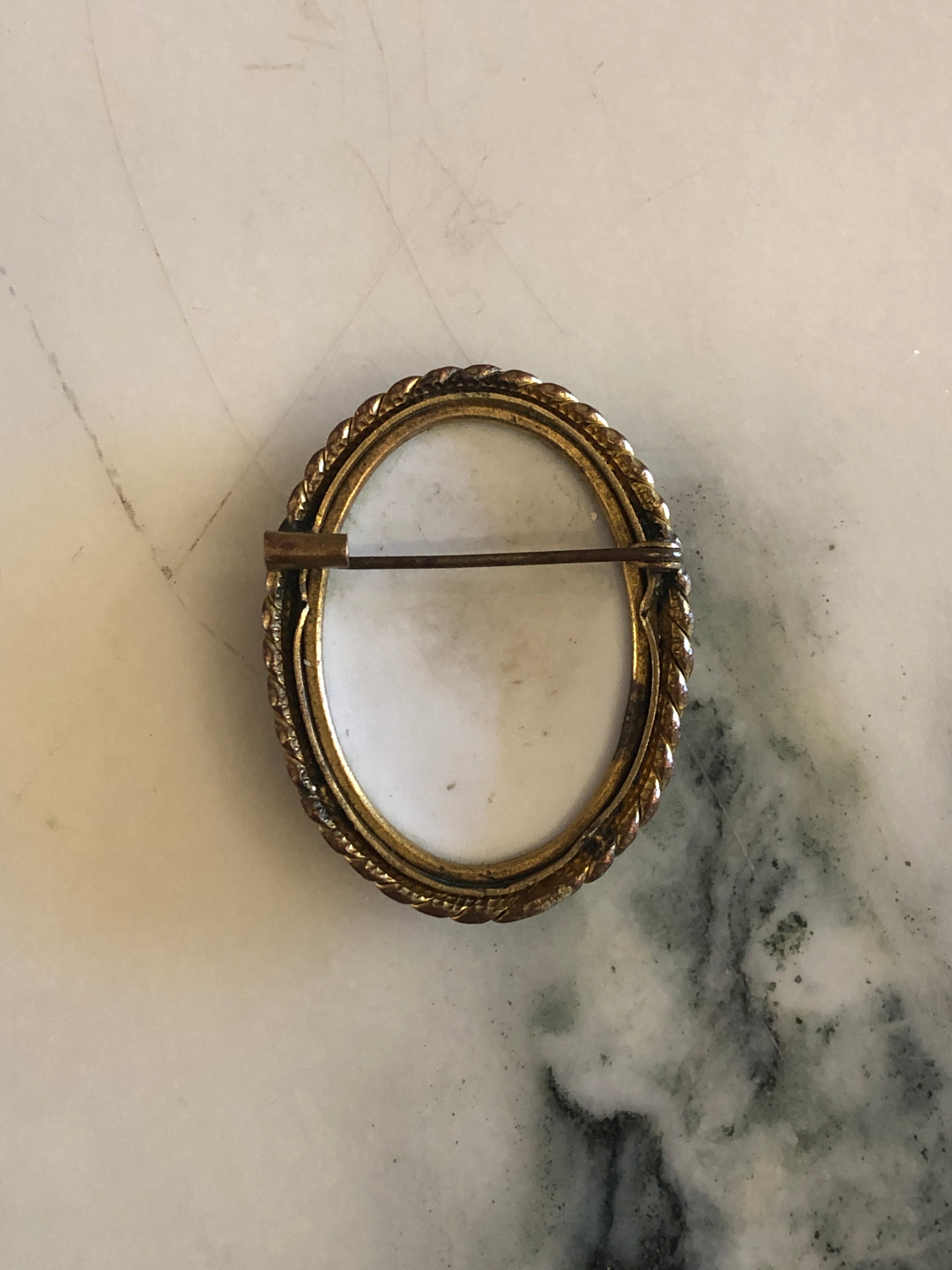 A 4cm x 3.5cm vintage cameo brooch with a carved white veiled classical portrait on a deep green ground in a gold-tone twisted rope frame, photographed on a white marble surface.