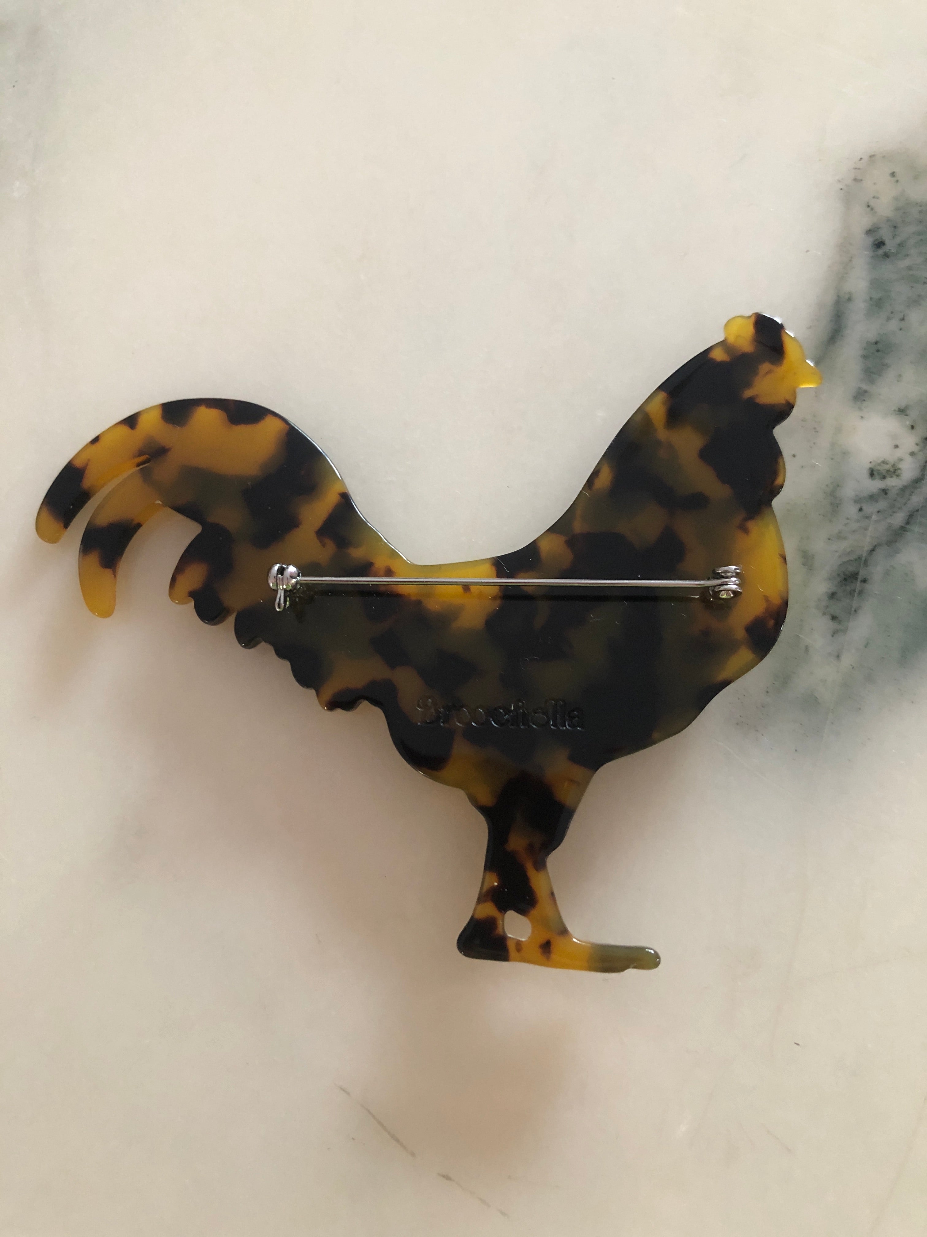 A Broochella Animalia Collection beige tortoiseshell rooster brooch and men's lapel pin in Italian cellulose acetate. The background is white marble