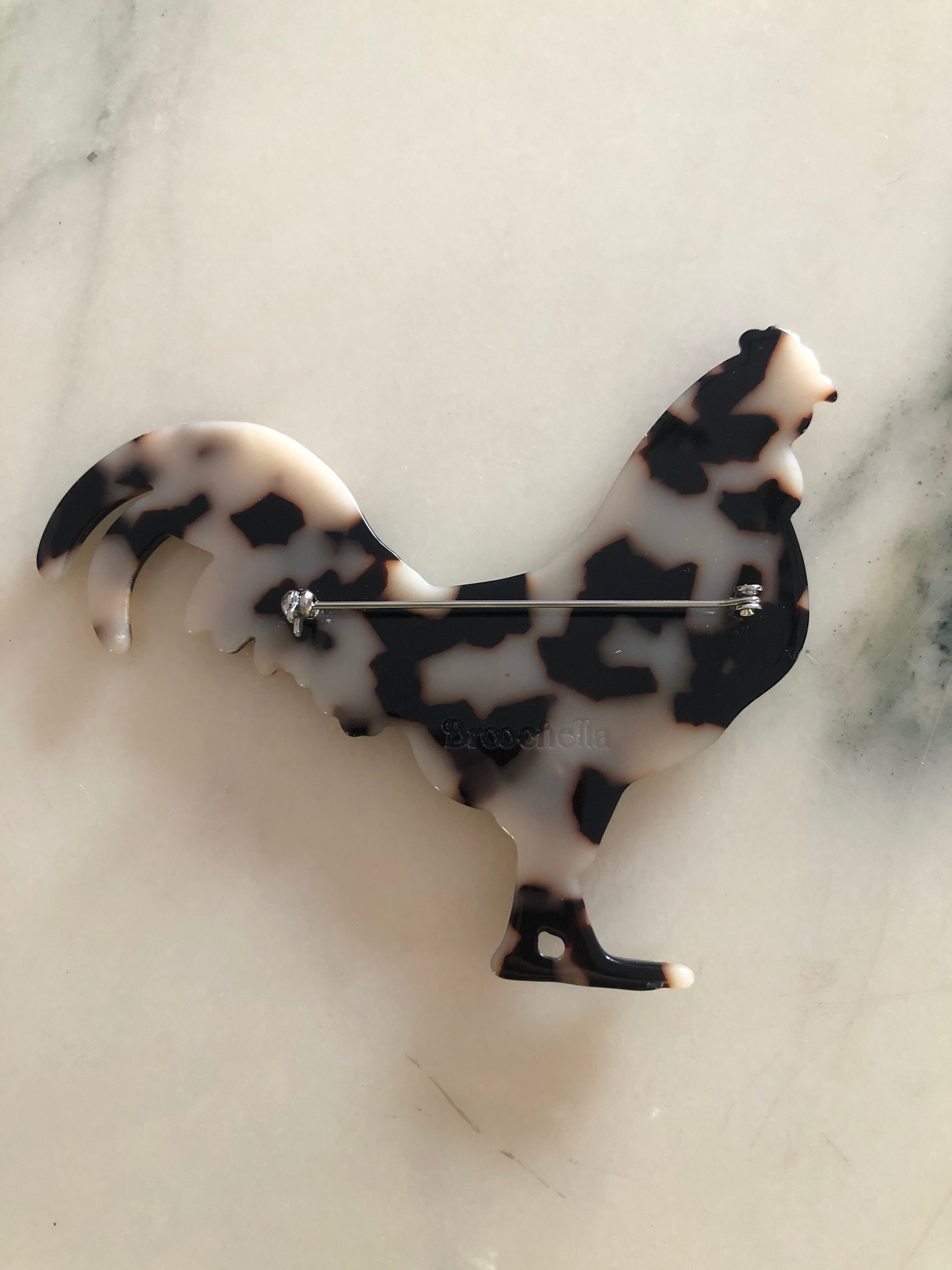 A Broochella Animalia Collection rooster brooch in brown and amber tortoiseshell Italian cellulose acetate with sculpted feather and tail detail measuring 8cm x 10cm, photographed on a white marble surface.
