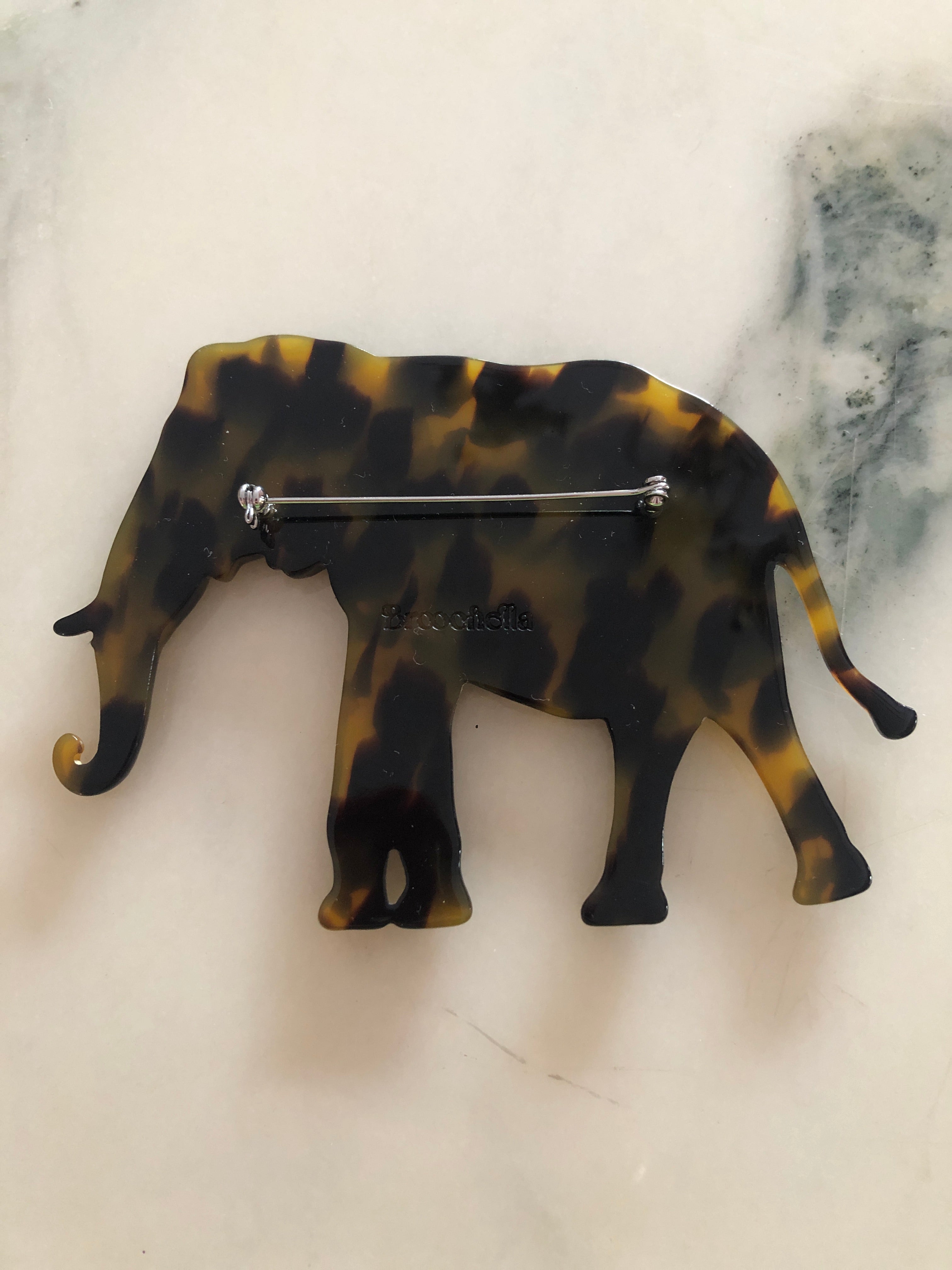 A Broochella Animalia Collection elephant brooch in black and white tortoiseshell Italian cellulose acetate with sculpted curled trunk detail measuring 8cm x 11cm, photographed on a white marble surface.