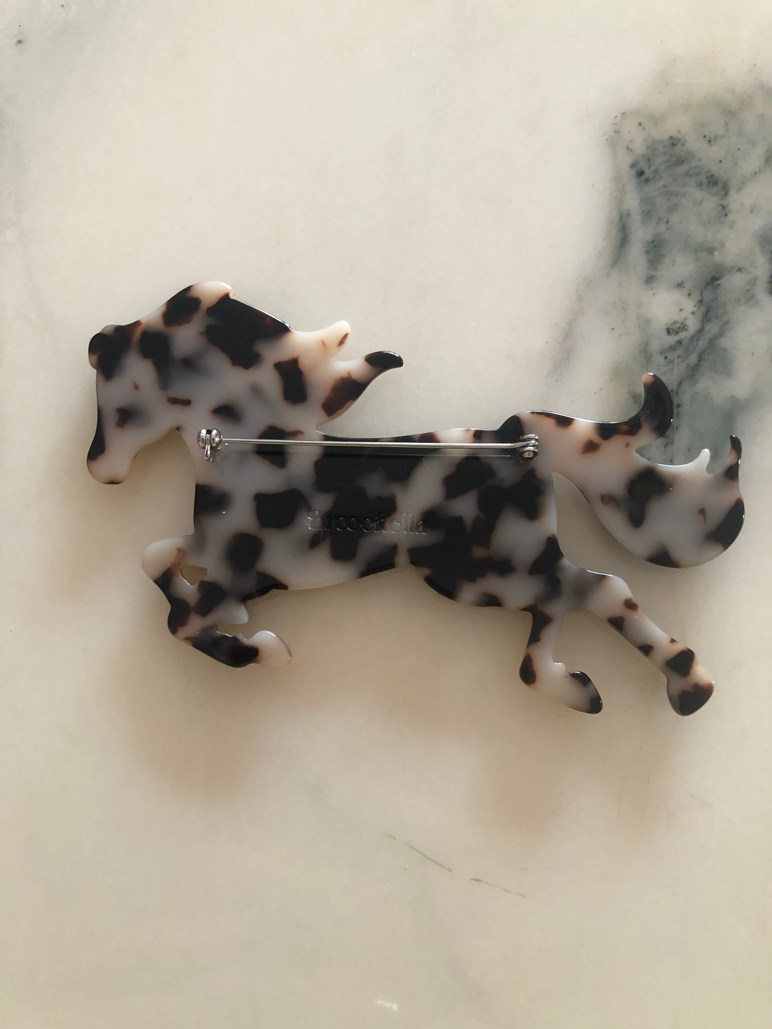 A Broochella Animalia Collection tortoiseshell running horse brooch in brown Italian cellulose acetate. The background is white marble