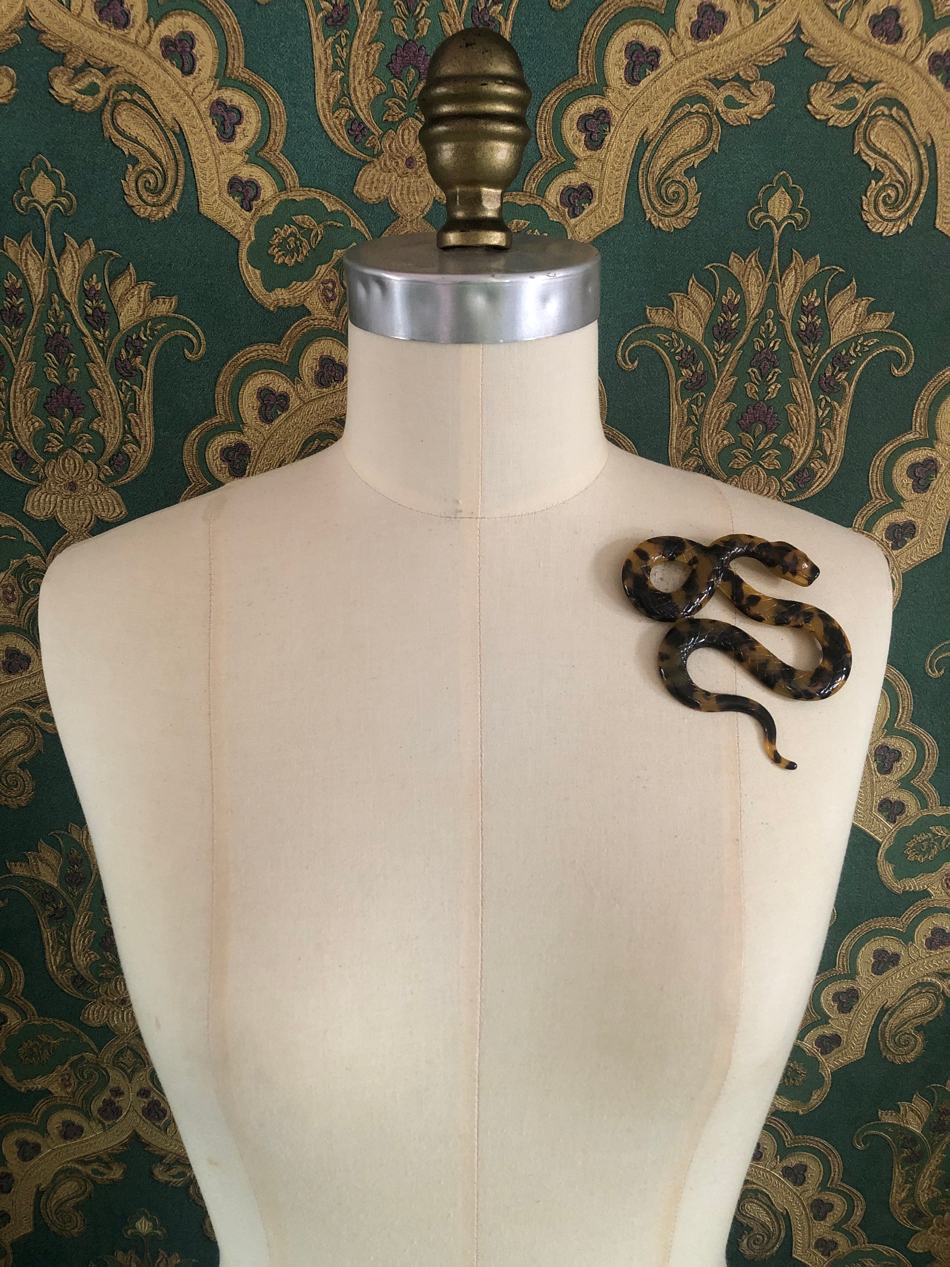 A Broochella Animalia Collection coiled snake brooch in black and white tortoiseshell Italian cellulose acetate with detailed scale texture measuring 9cm x 10cm, photographed on a white marble surface.