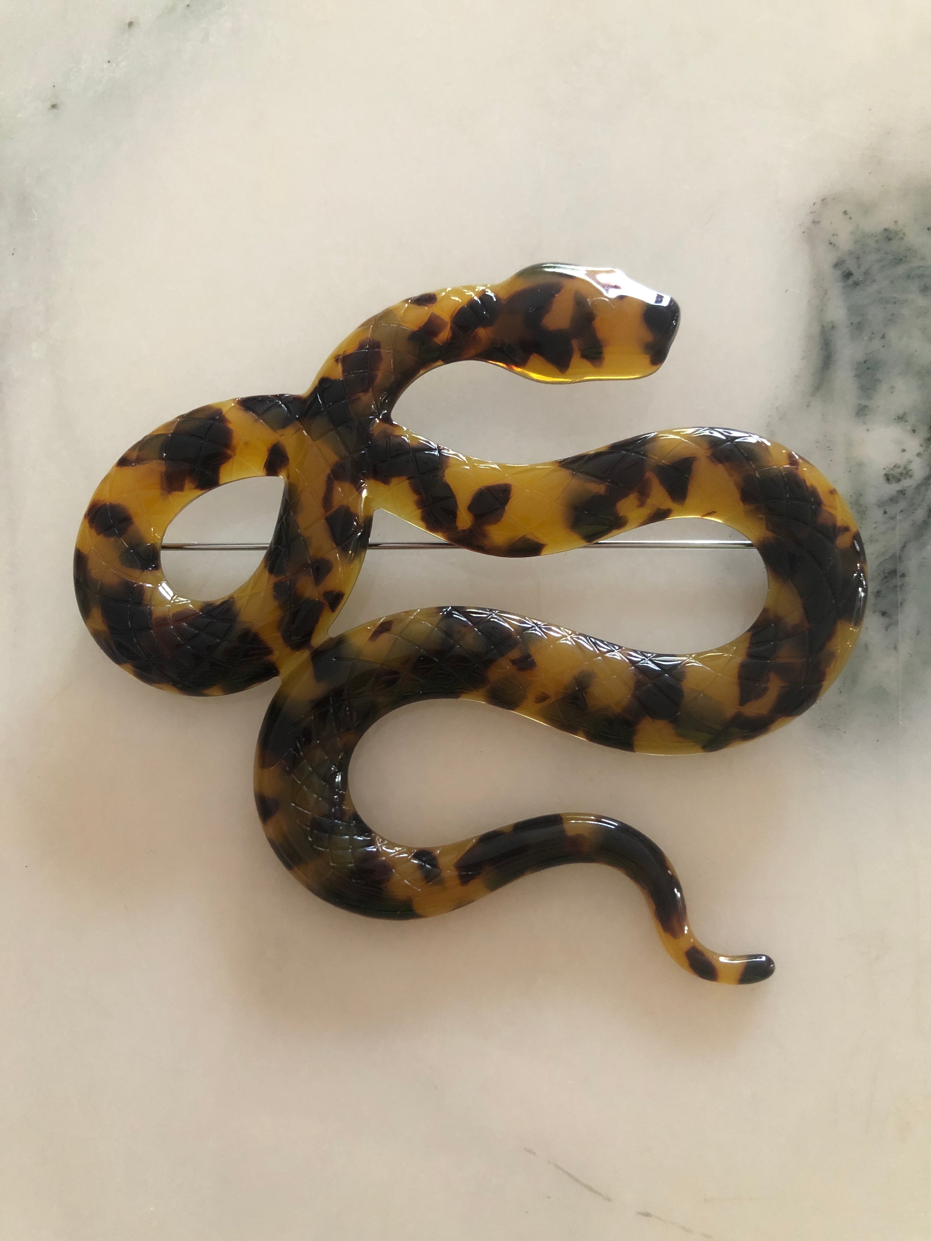 A Broochella Animalia Collection coiled snake brooch in black and white tortoiseshell Italian cellulose acetate with detailed scale texture measuring 9cm x 10cm, photographed on a white marble surface.
