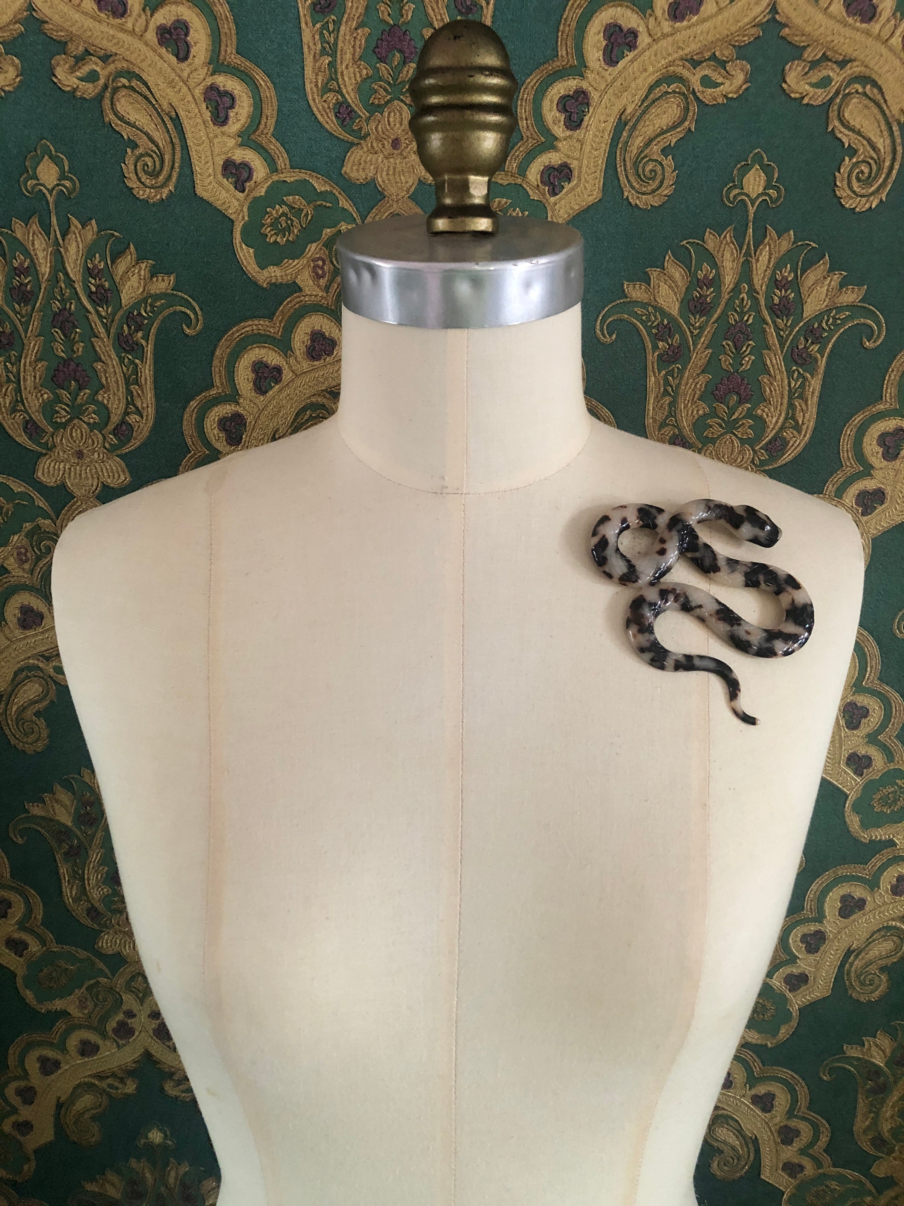 A Broochella Animalia Collection coiled snake brooch in black and white tortoiseshell Italian cellulose acetate with detailed scale texture measuring 9cm x 10cm, photographed on a white marble surface.